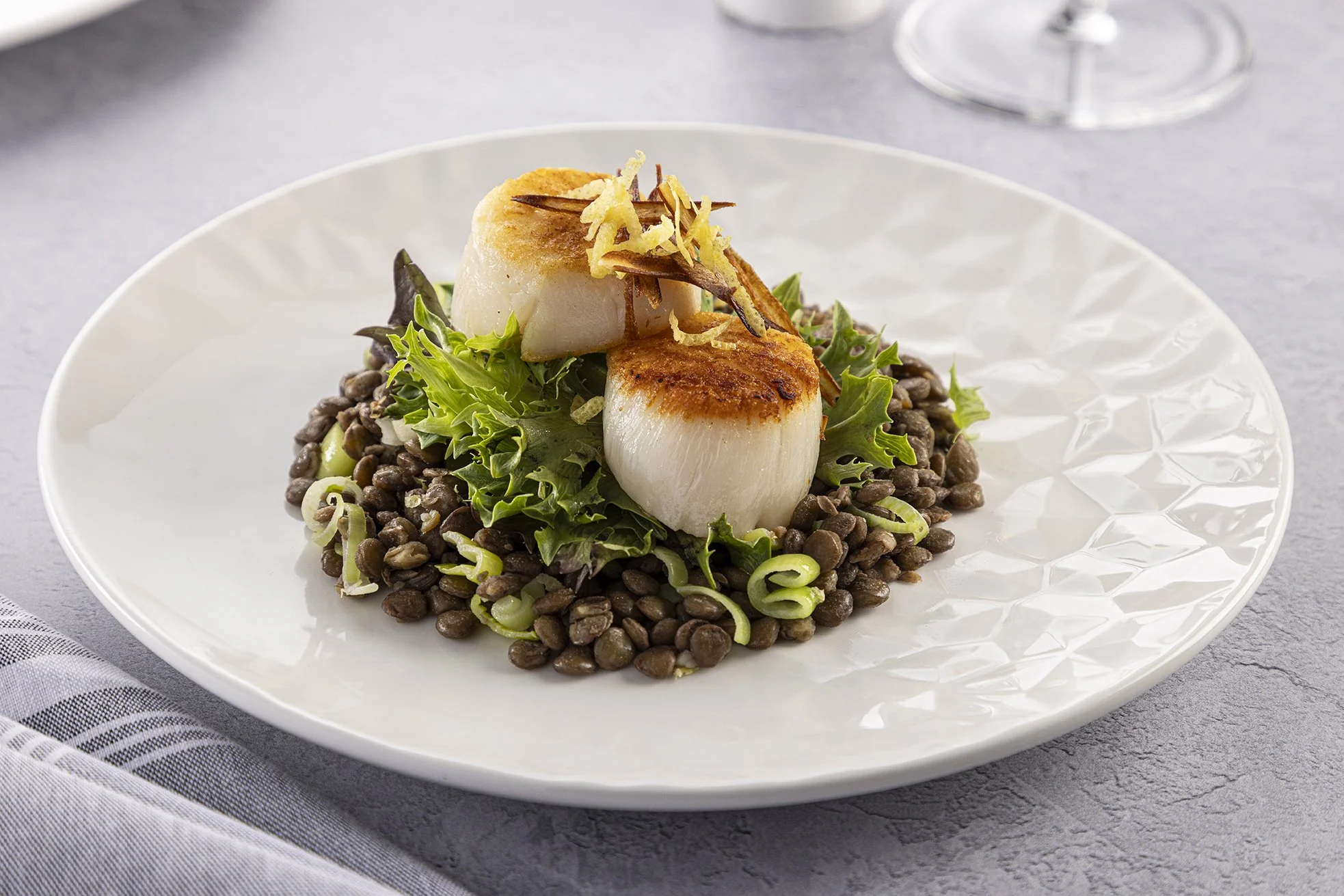 Seared scallops served over black lentils, garnished with crispy fried shallots and herbs on a white plate