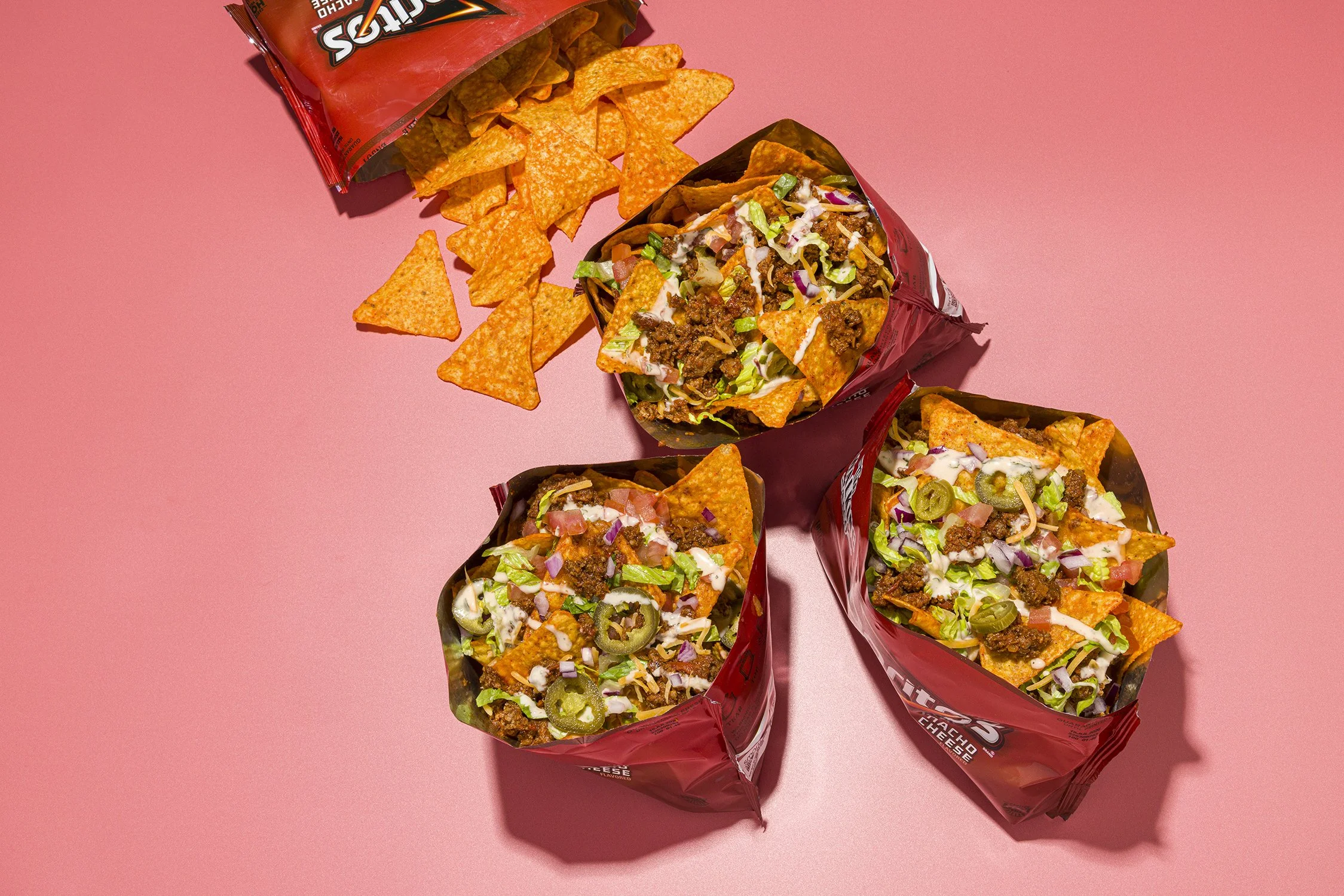 Five Taco Bell Cheddar Cheesy Gordita Crunch wraps with nacho cheese, lettuce, tomatoes, jalapenos, sour cream, and ground beef, along with a scattered pile of Doritos Flamin' Hot Nacho cheese Doritos chips on a pink background.