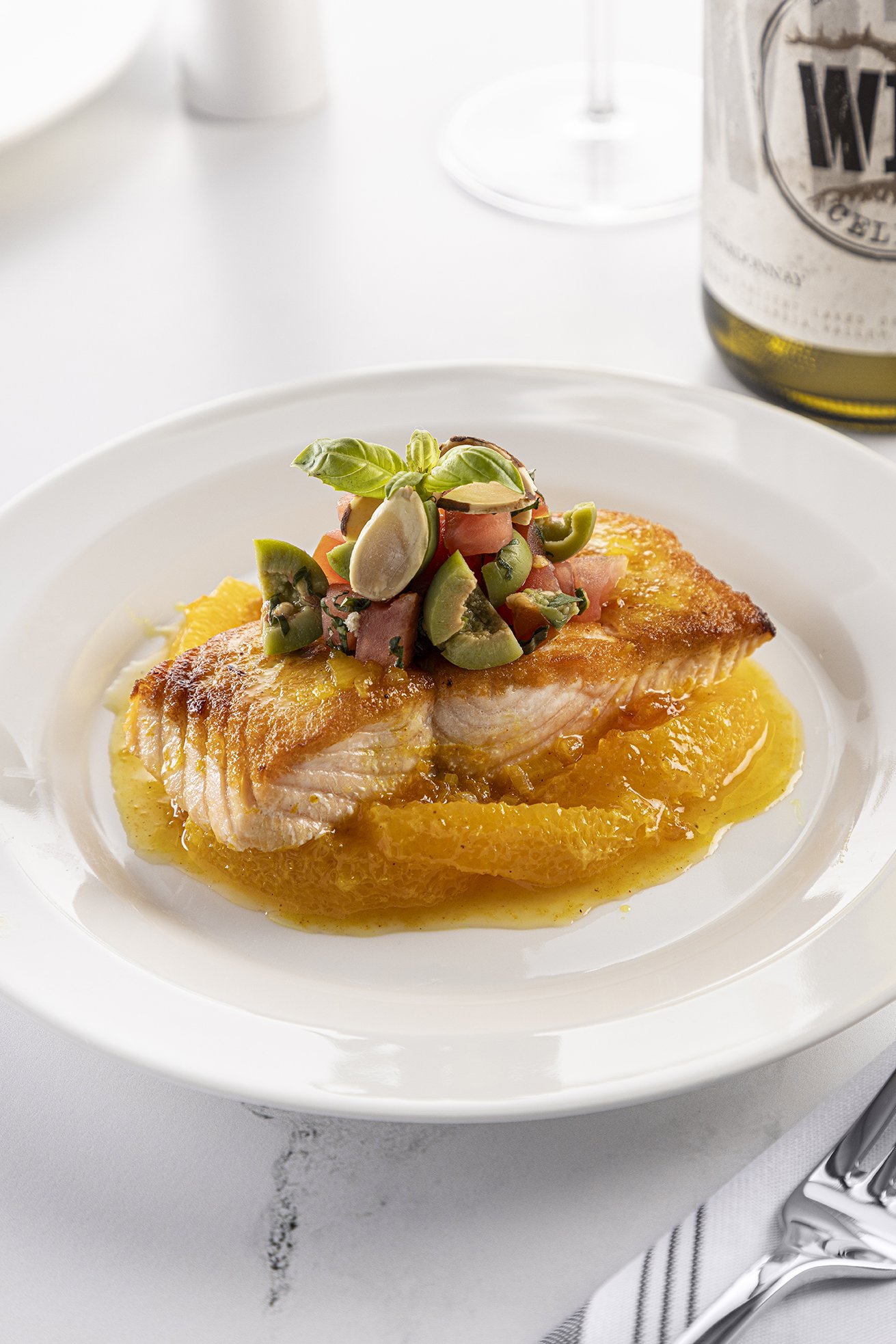 A plated dish with a cooked fish fillet topped with chopped vegetables and herbs, served with a yellow sauce on a white plate, with a bottle of wine and a glass in the background.