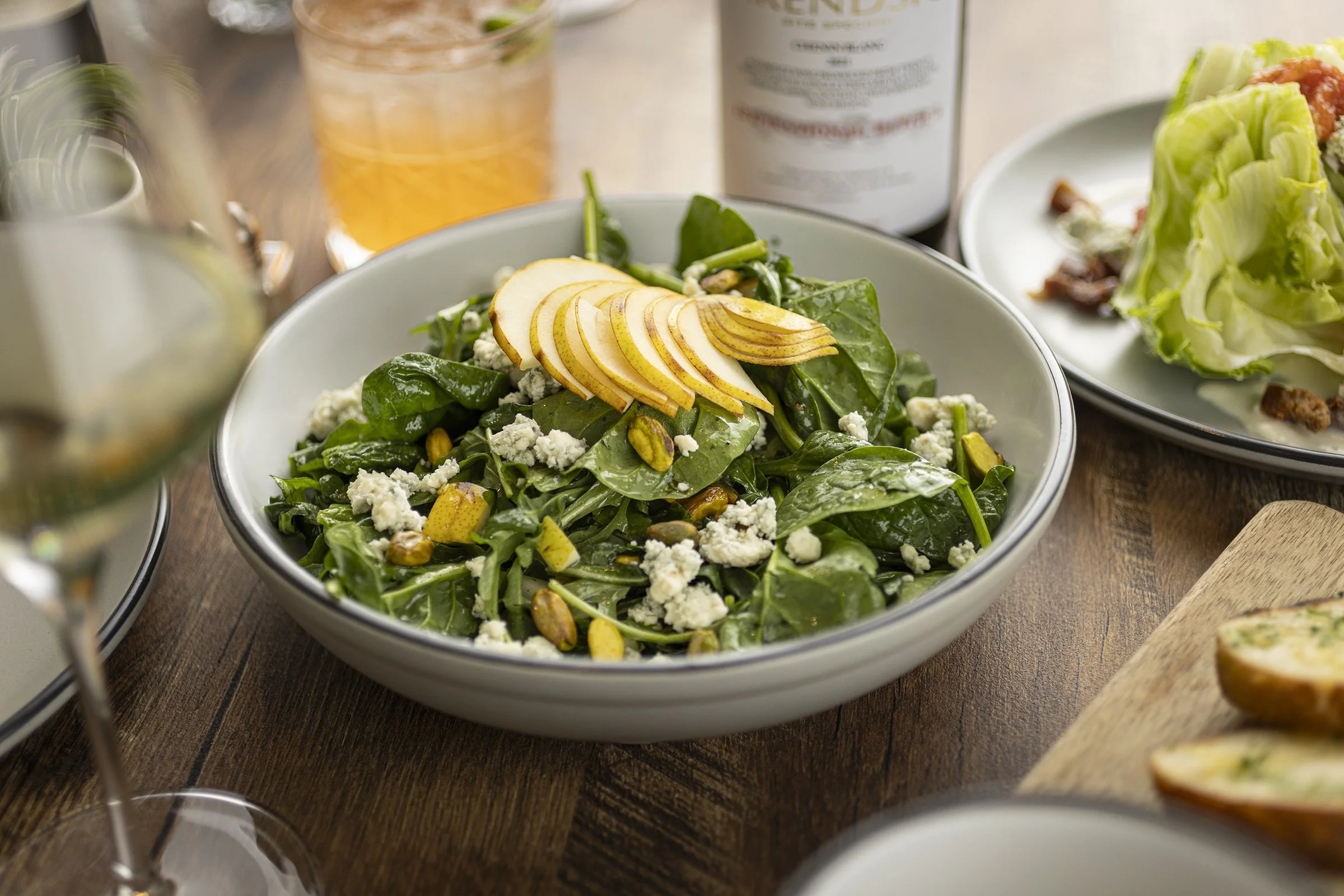 A bowl of spinach salad topped with sliced pears, crumbled blue cheese, and pistachios, with a glass of iced drink and a bottle in the background.