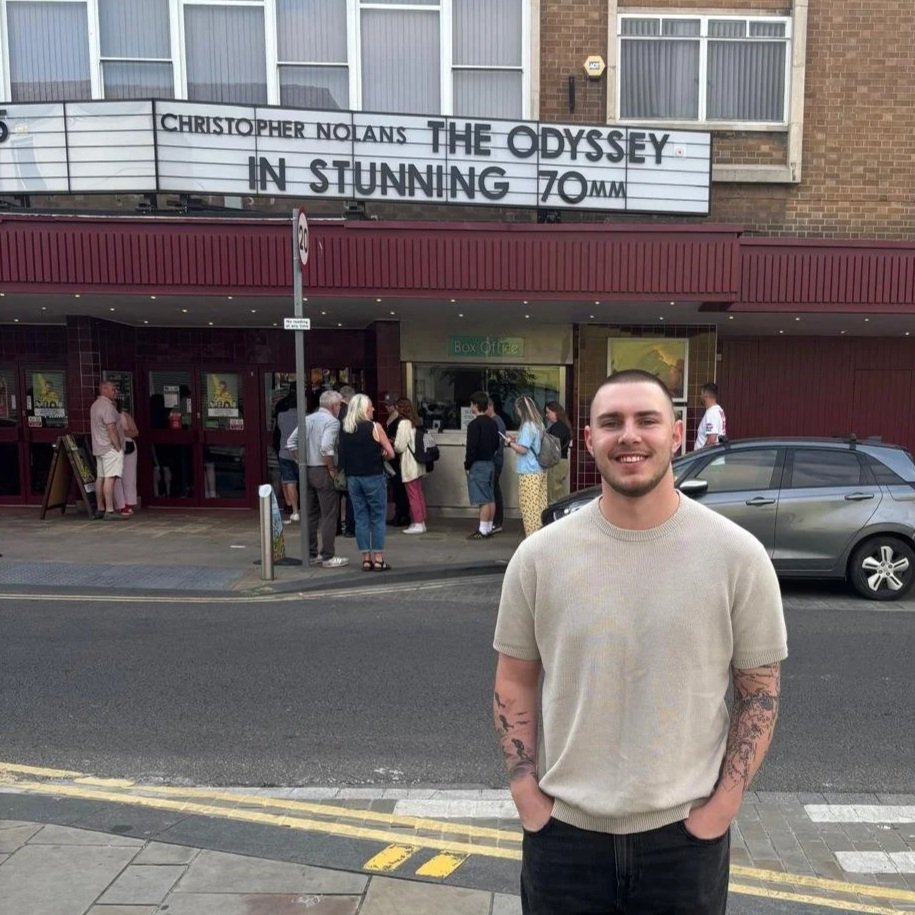 Young man with tattoos standing on a city street in front of a theater, smiling, with people lined up outside the box office, and a marquee advertising Christopher Nolan's film "Oppenheimer" in 70mm.