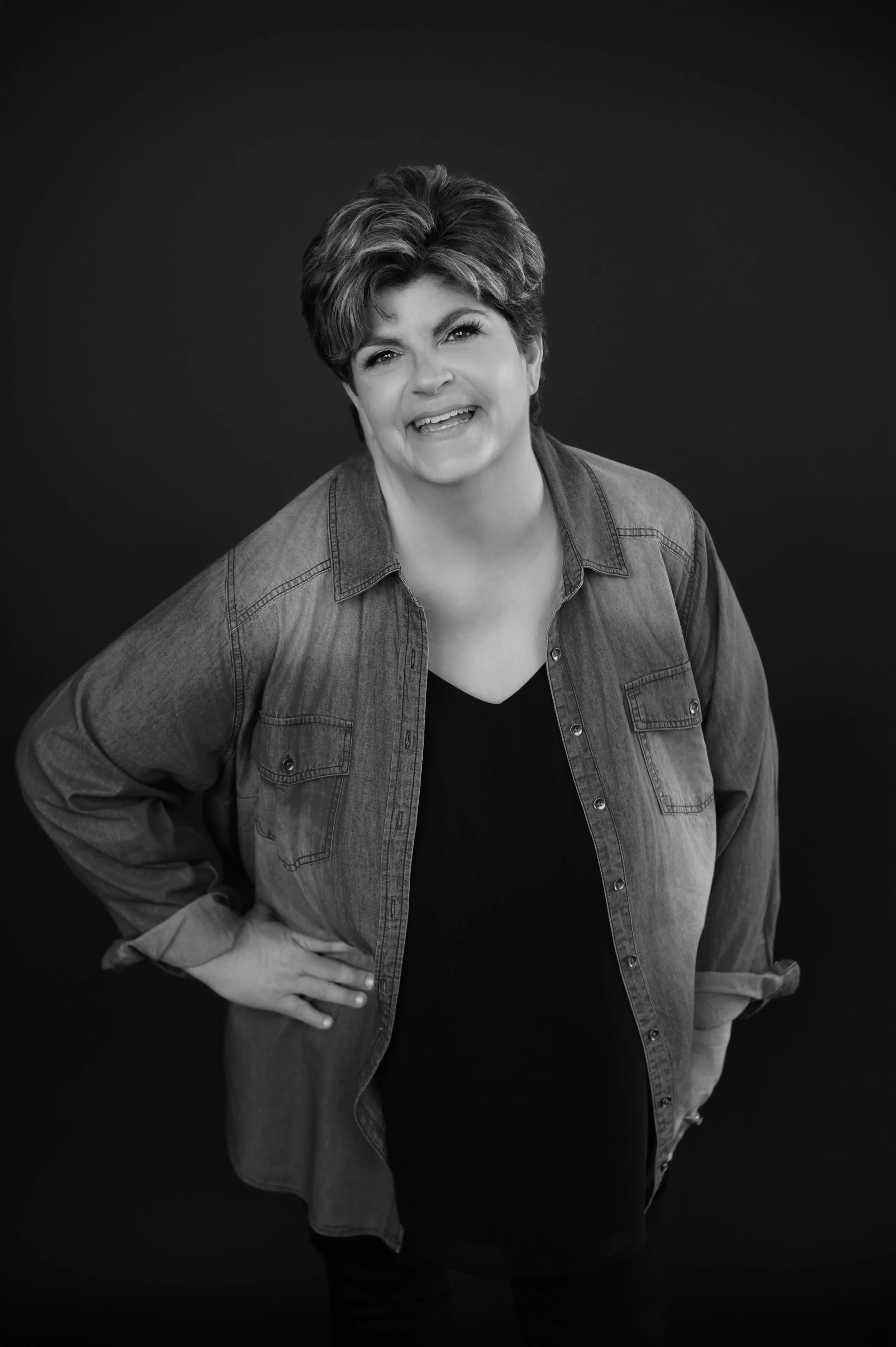 Black and white portrait of a woman with short hair, wearing a denim shirt over a dark top, smiling and with her hands on her hips, standing against a dark background.