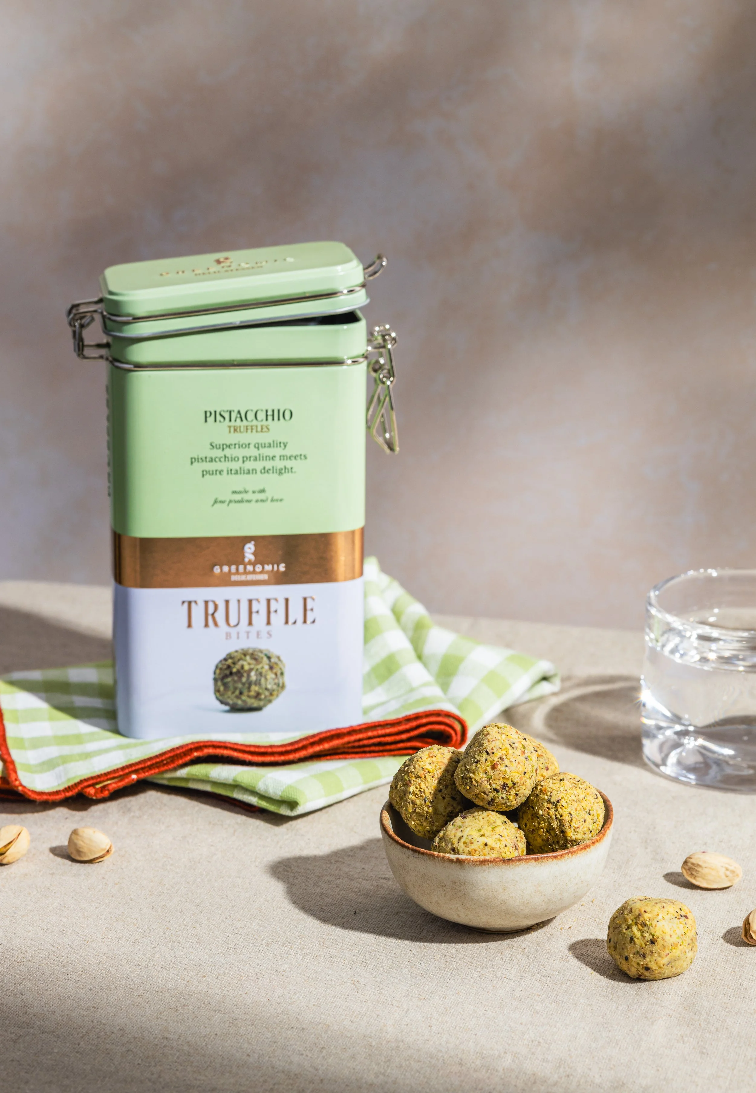 Eine offene Blechdose mit grüner Oberfläche, die Pistazien-Trüffel enthält, auf einem Gedeck in der Mitte. Daneben steht eine kleine Schale mit Pistazien-Trüffeln. Auf einer grün-weiß karierten Serviette liegt ein rot gerändeltes Tuch. Ein Sektglas m