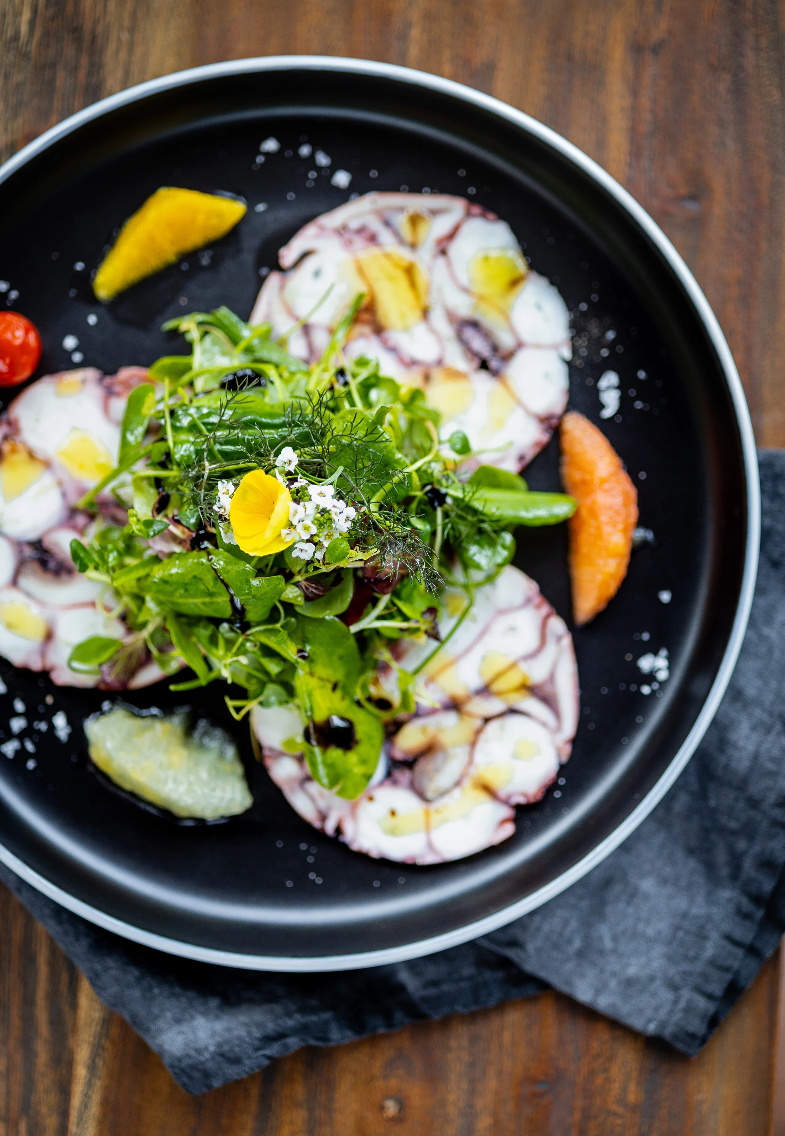Carpaccio vom mediterranen Oktopus mit Zitrusfrüchten auf einem dunklen Teller.