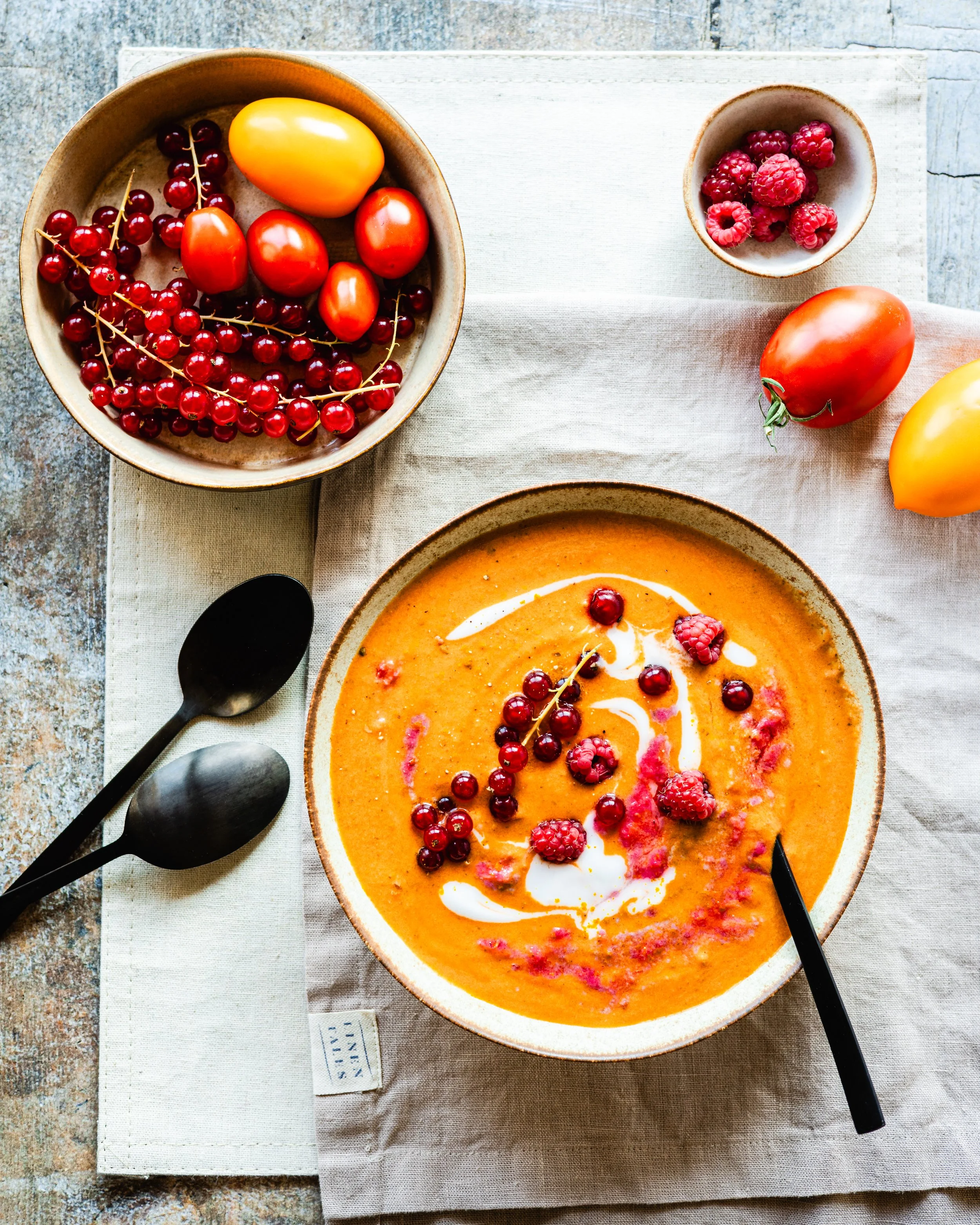 Ein Teller mit einer cremigen orangen Suppe, garniert mit roten Johannisbeeren, Himbeeren und einem Schuss Sahne, daneben ein Löffel. Im Hintergrund befindet sich eine Schüssel mit Johannisbeeren, sowie Tomaten auf einem Tuch auf einem Holztisch.