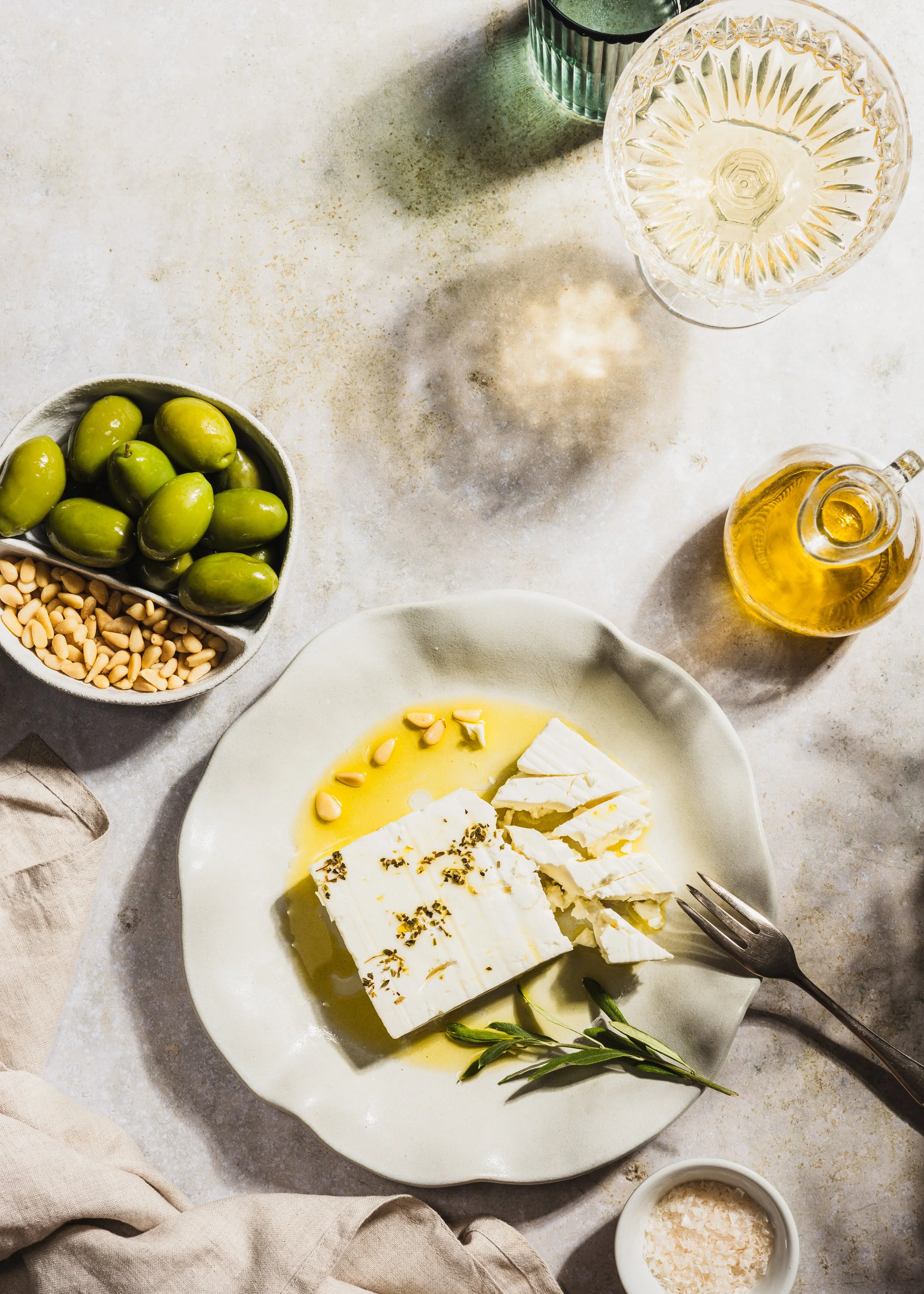eine Platte mit Feta-Käse, garnierte mit schwarzen Pfeffer und Rosmarin, daneben eine kleine Schüssel mit Meersalz, ein Glas mit weißem Wein, eine Schüssel mit grünen Oliven, Pinienkerne in einer kleinen Schale, und eine Trinkflasche mit Öl. Ein Mess