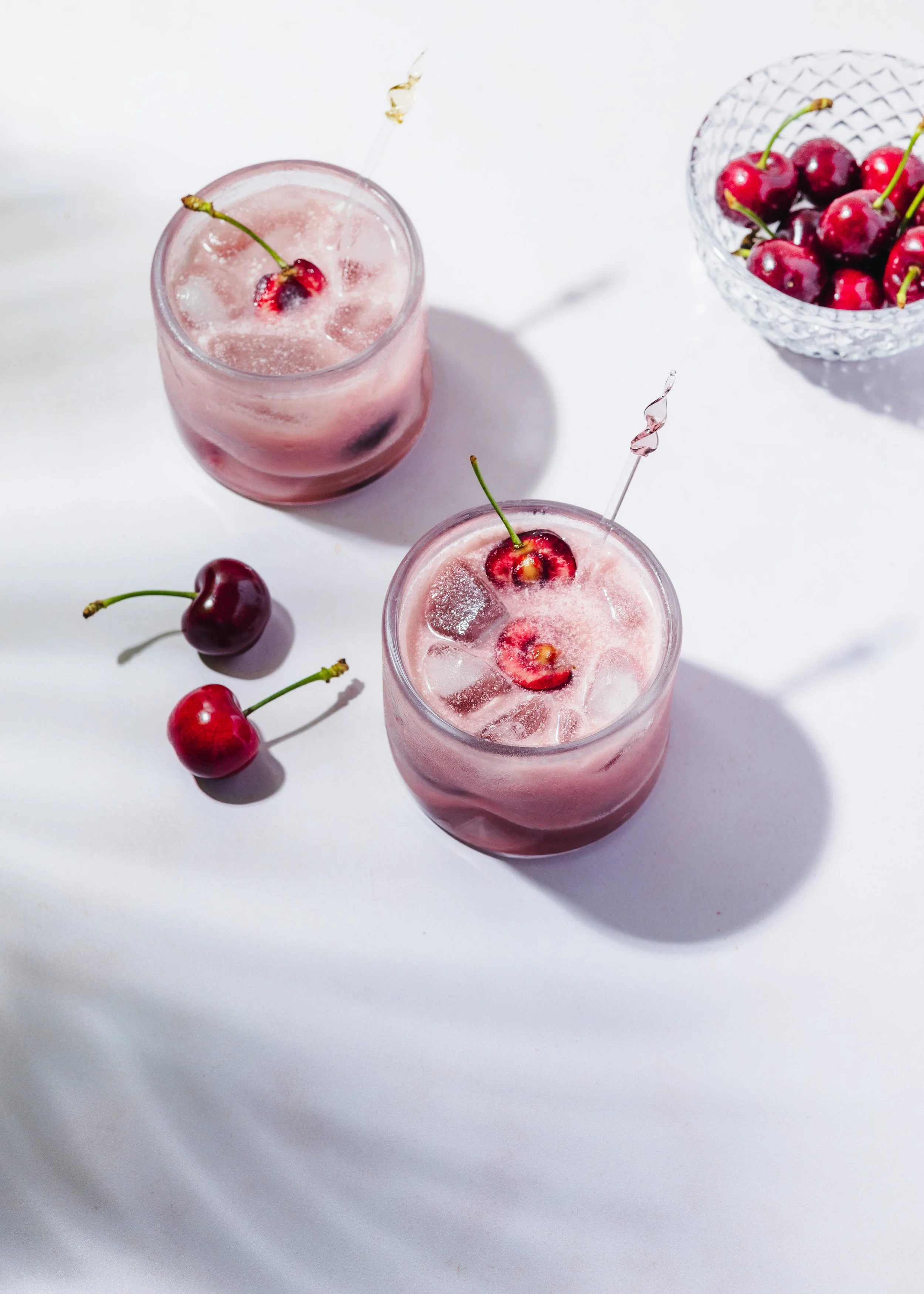 Zwei Gläser mit rosa Getränk und Kirschen, ein Kristall-Schüssel mit Kirschen, auf weißem Hintergrund.