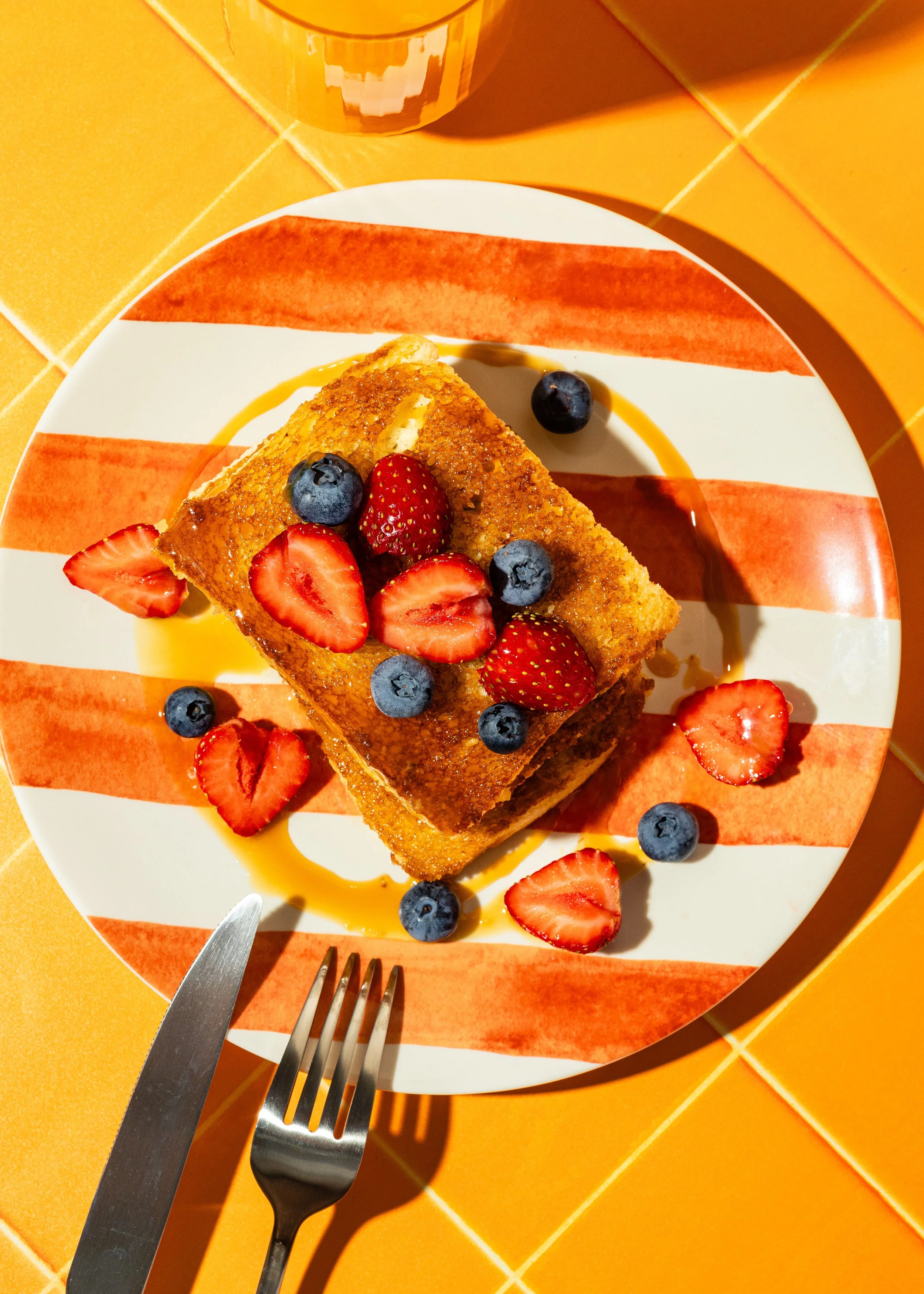 Ein Teller mit einem Stück Pfannkuchen, garniert mit frischen Erdbeeren, Heidelbeeren und einem Schuss Sirup, auf einem farbenfrohen, gestreiften Teller, auf einem orangefarbenen Tisch.