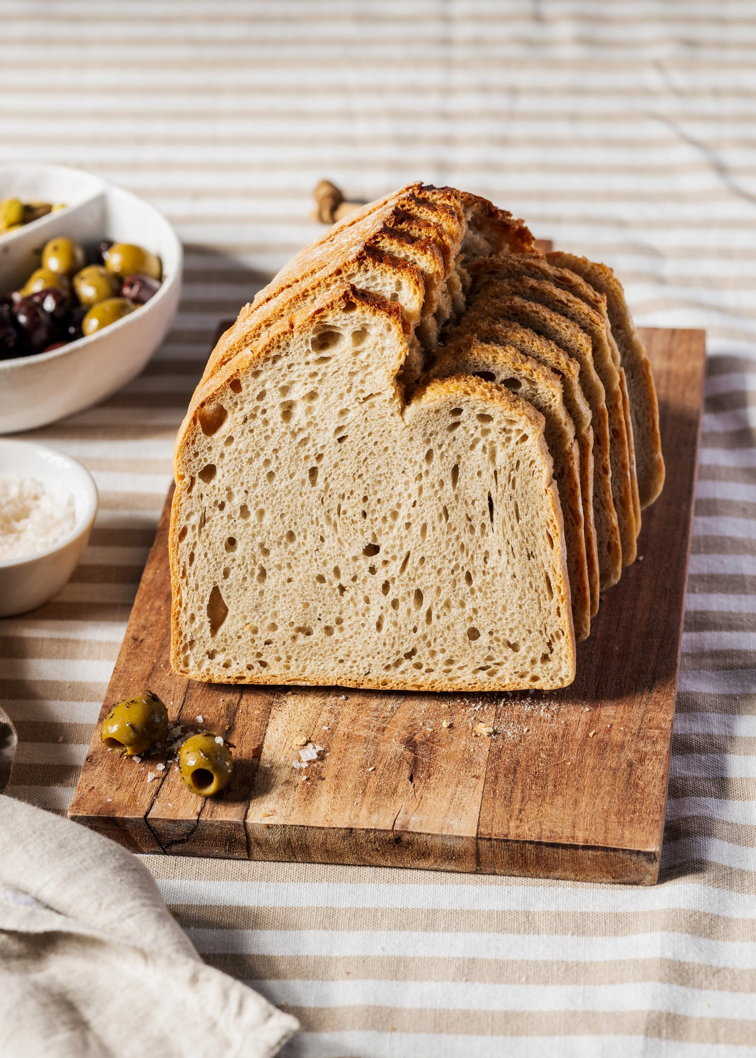 Scheibe hausgebackenes Brot auf einem Holzbrett, im Hintergrund eine Schüssel mit grünen und schwarzen Oliven, daneben kleine Schälchen Salz, und eine Stoffserviette