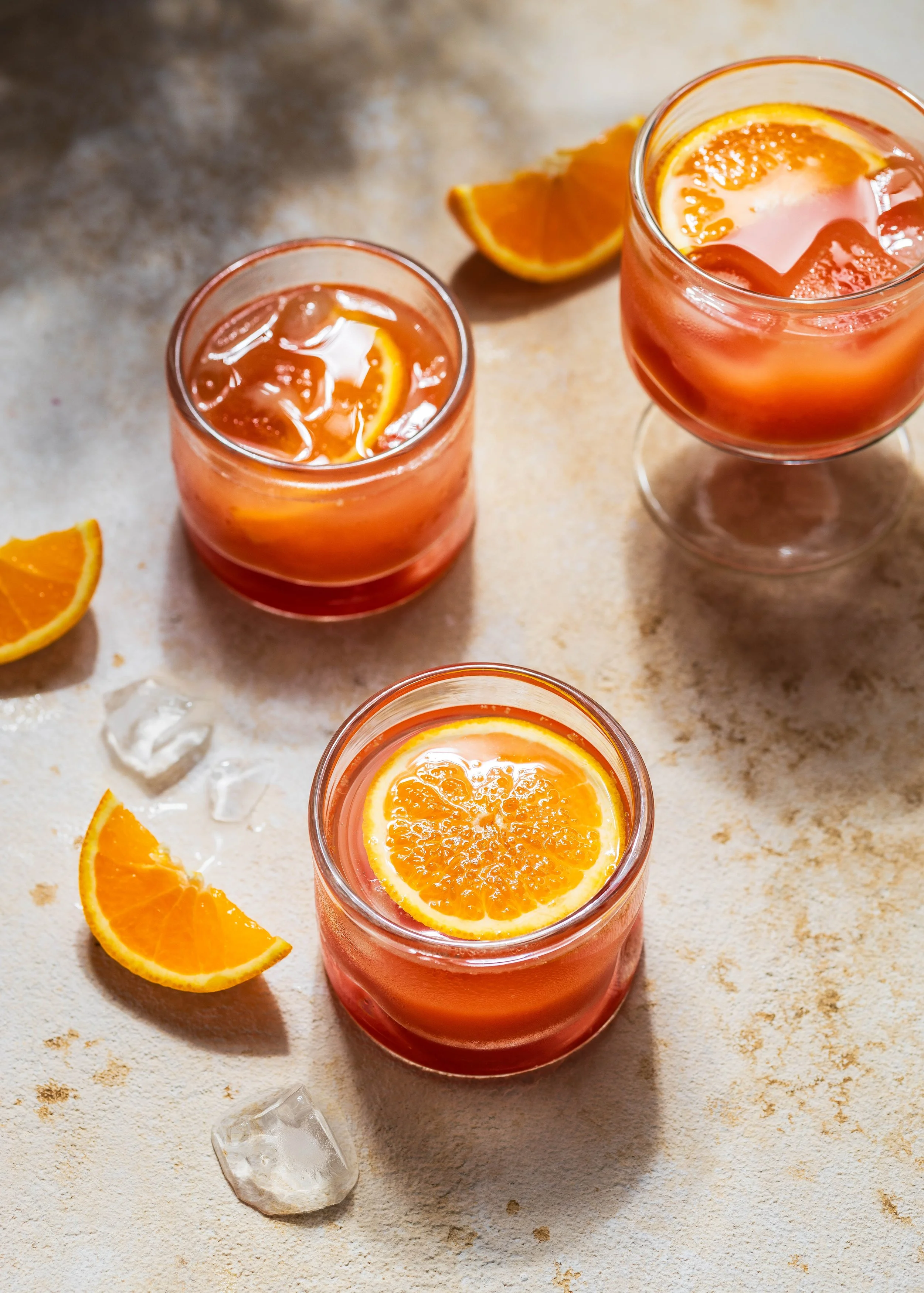 Drei Gläser mit Aperol Spritz und Orangenscheiben, Zitronenscheiben und Eiswürfel auf einem hellen, strukturierten Tisch.
