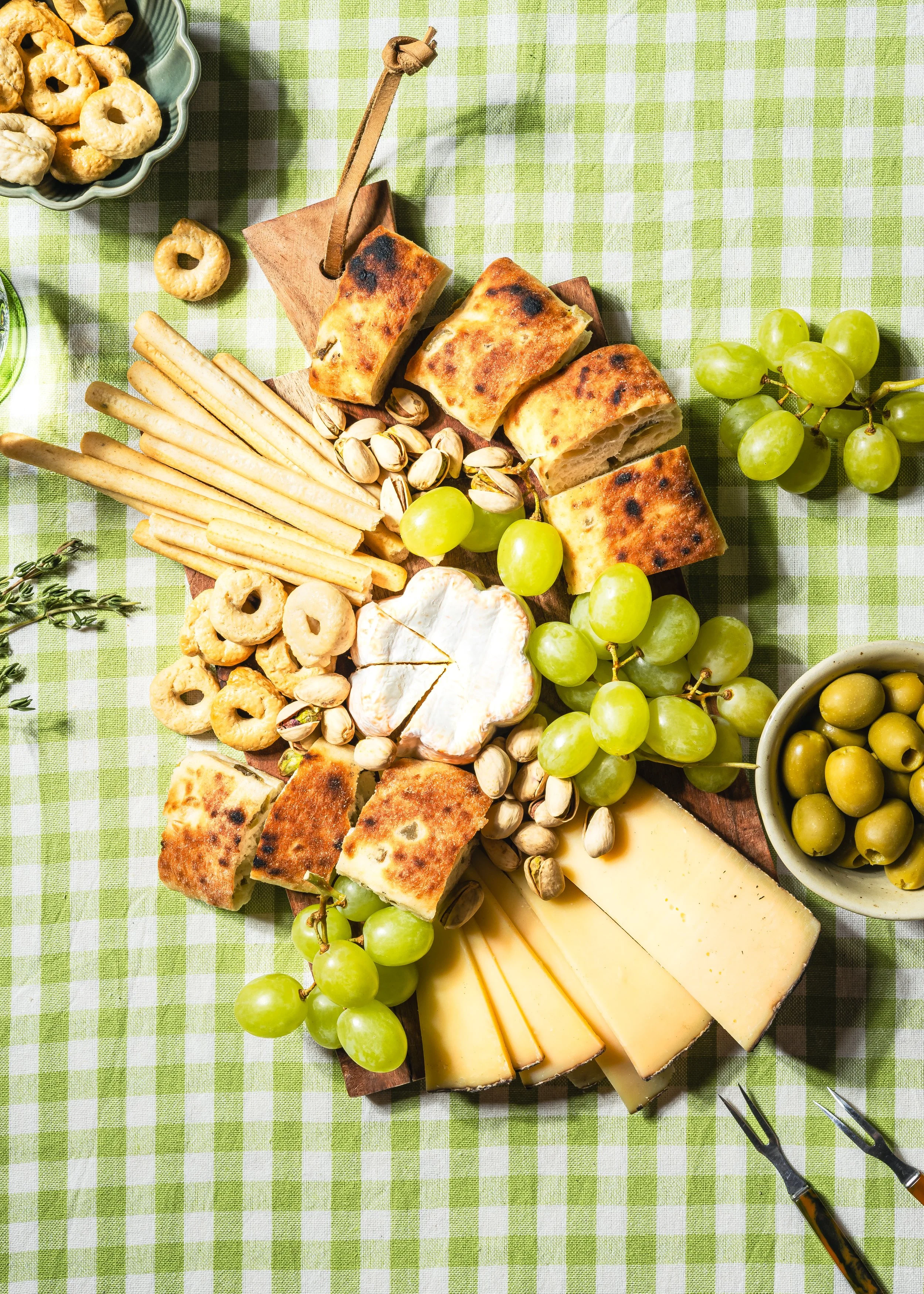 Käse, Brot, Trauben, Oliven, Pistazien, Knabbergebäck und Kräuter auf einem grünen Karoboden.