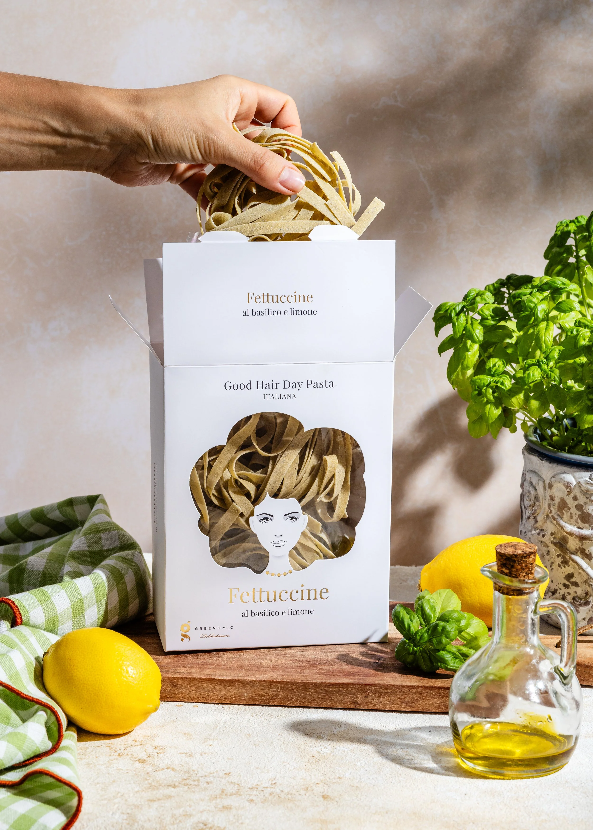 Schachtel mit veganen Fettuccine Nudeln mit Basilikum und Zitrone, umgeben von frischen Zitronen, Basilikum und einem Flasche Olivenöl, auf einem Holztisch mit grün-weiß karierter Stoffserviette.