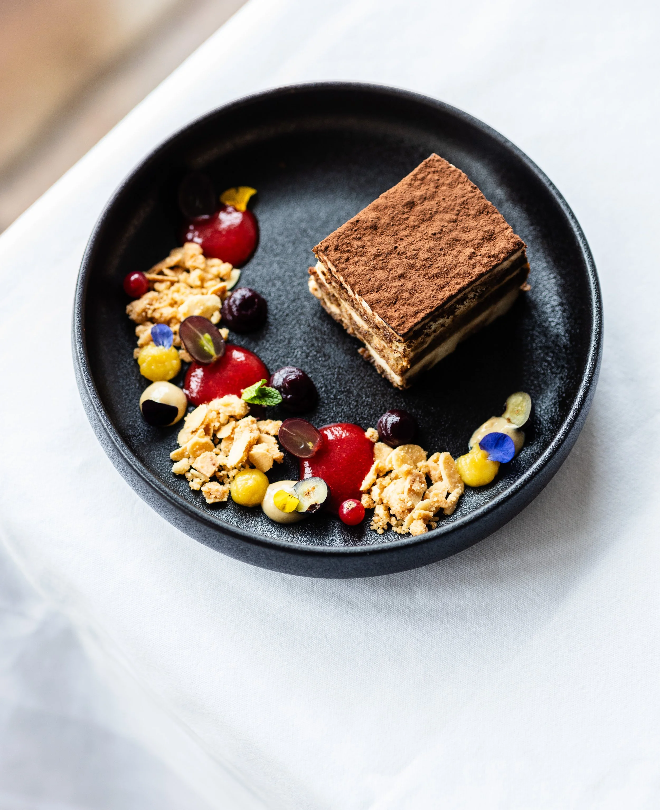 Ein Stück Tiramisu auf einem schwarzen Teller, dekoriert mit Beeren, Nüssen, Blüten und Cremetupfen.