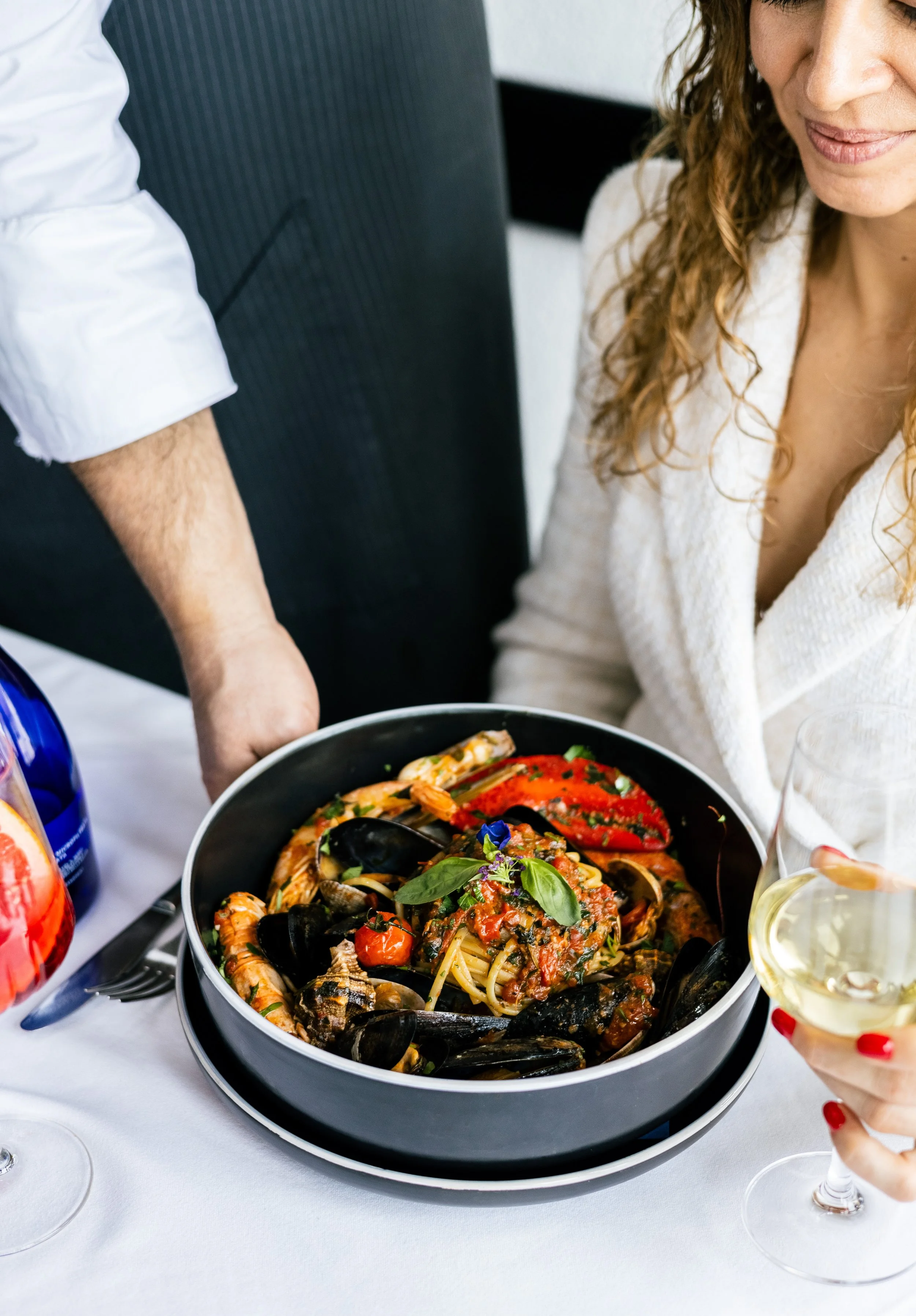 Ein großer Topf mit Meeresfrüchtespi spaghetti, garniert mit Basilikum und Blüten, auf einem festlichen Esstisch. Im Vordergrund ein Glas Weißwein, im Hintergrund eine Kellnermanschette, eine Flasche Wasser und eine Gabel. Eine Frau mit roten Fingern
