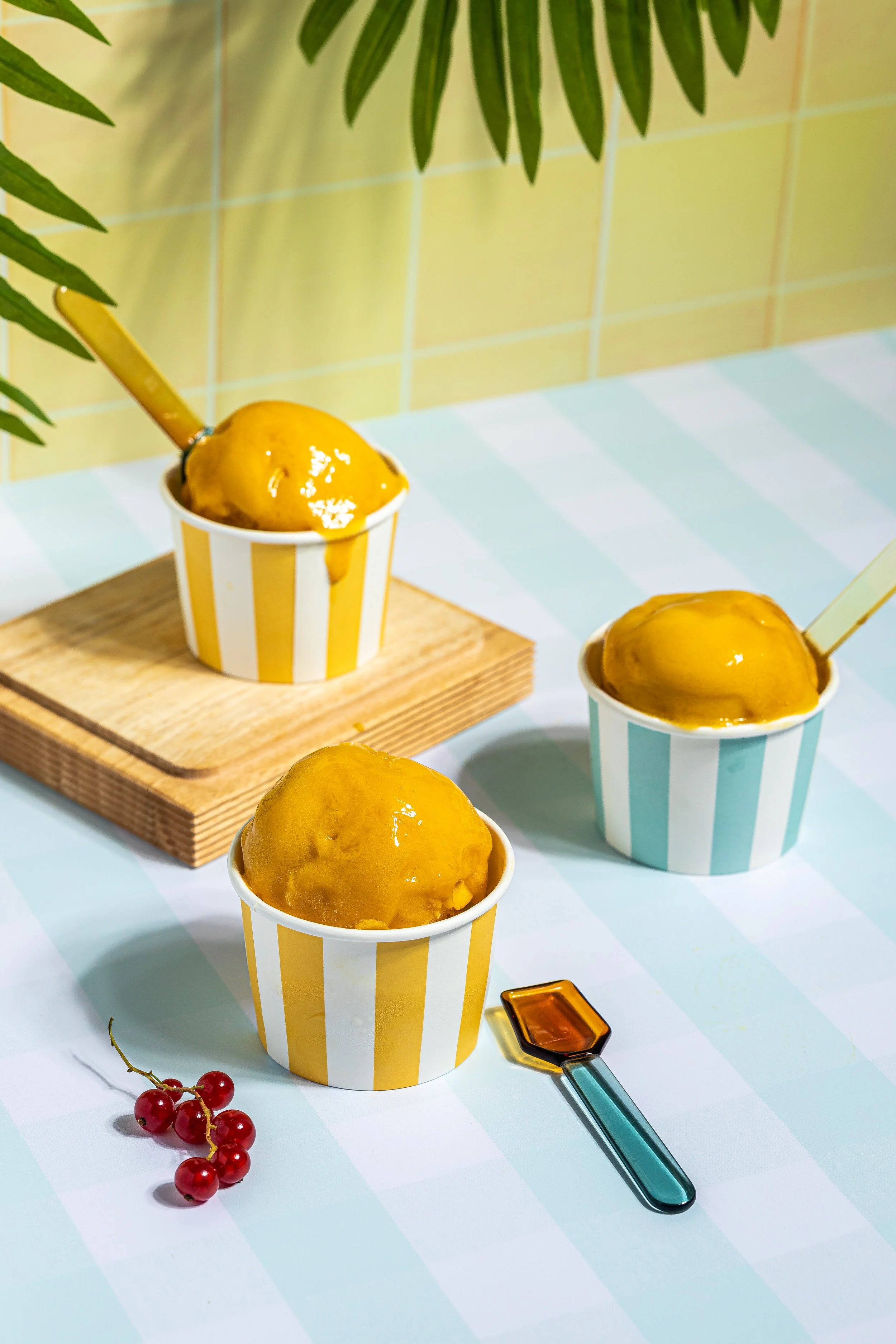 Erdbeeren und Vanilleeis in gelb-weiß gestreiften Bechern, mit einer Gelatospachtel neben einem Becher, auf einem weißen und blauen karierten Tisch, im Hintergrund grüne Blätter, Aufsteller auf Holzplatte.