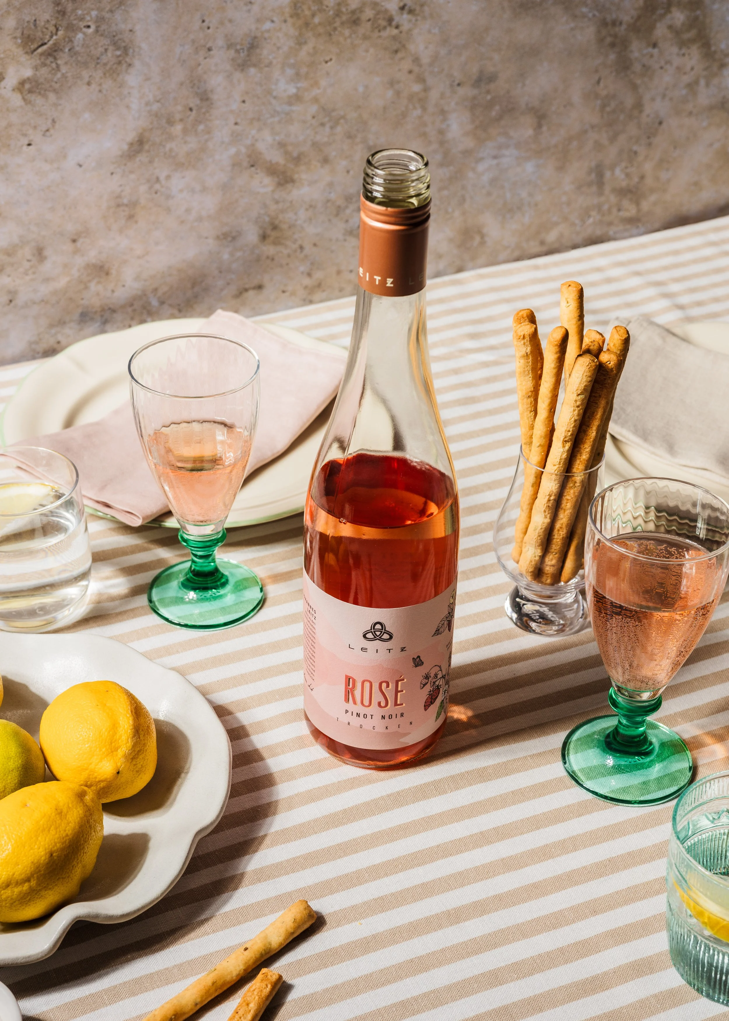 Bilder eines Esstisches mit einer Flasche Roséwein, Gläsern, einer Schale mit Zitronen und Keksen, sowie Grissini auf einem Tischdecken mit Streifenmuster.