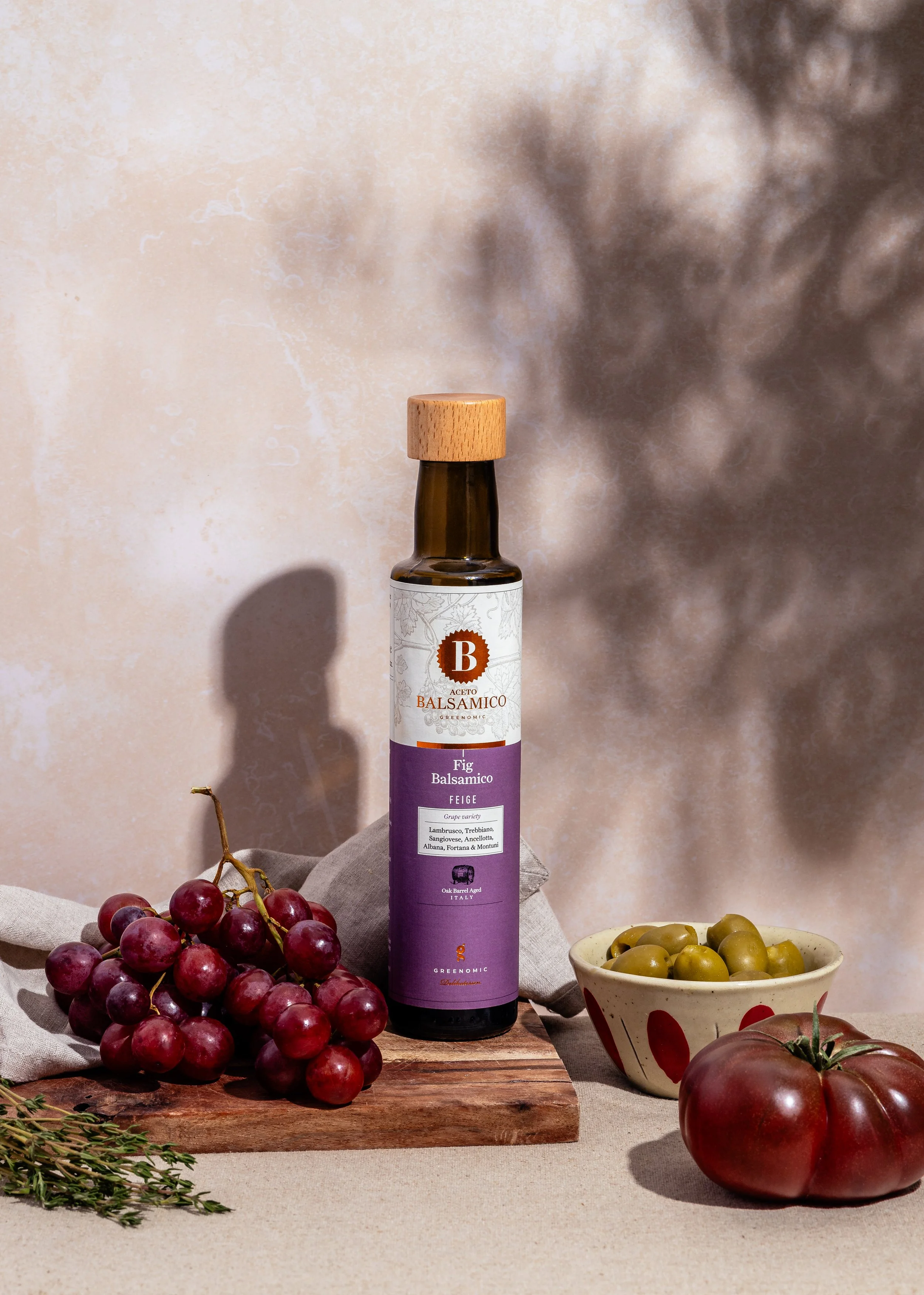 Ein Flasche Balsamico-Essig, umgeben von Trauben, Tomaten und einer Schale mit grünen Oliven auf einem Holzbrett