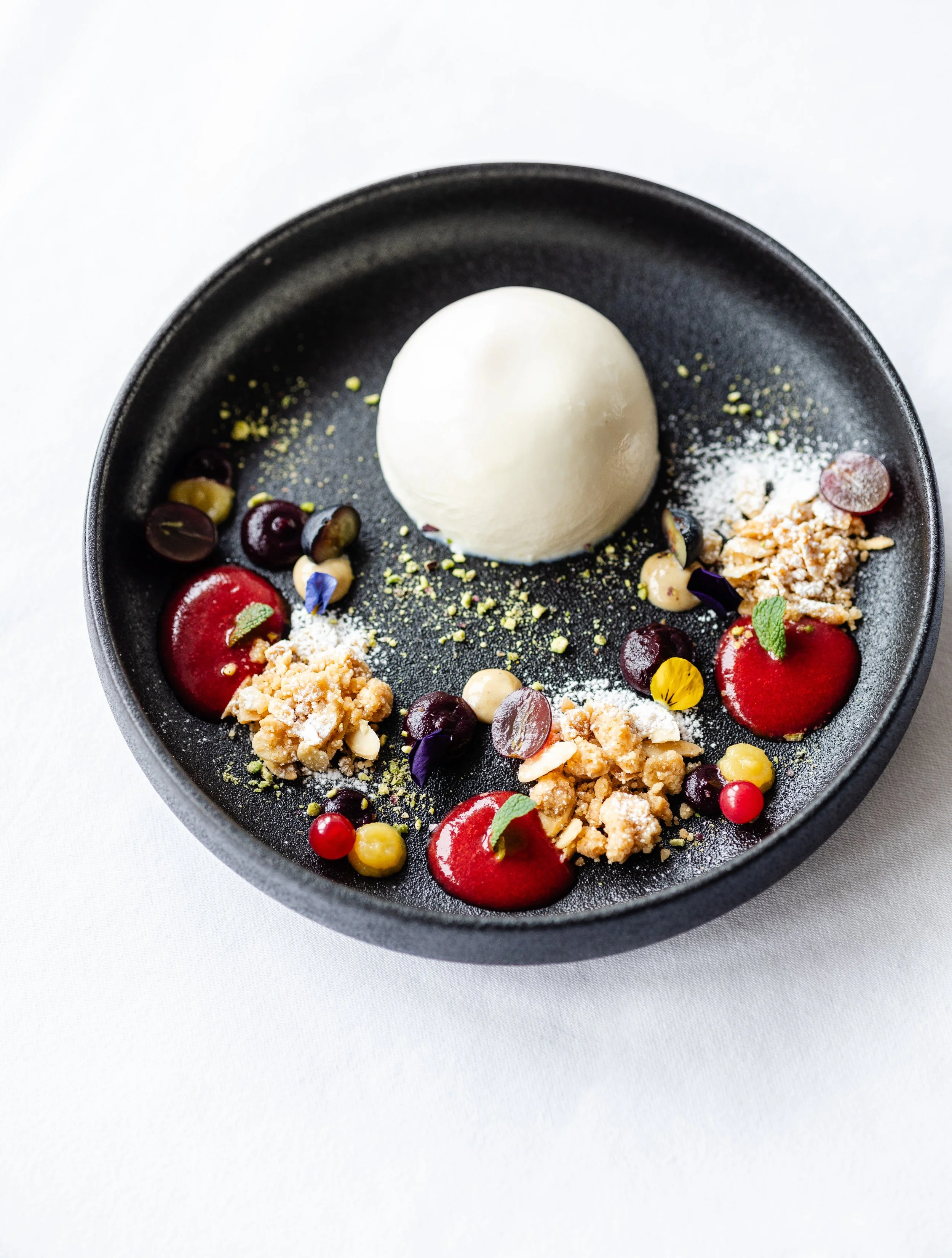 Panna Cotta mit Beeren, Nüssen und essbaren Blüten auf einem schwarzen Teller.