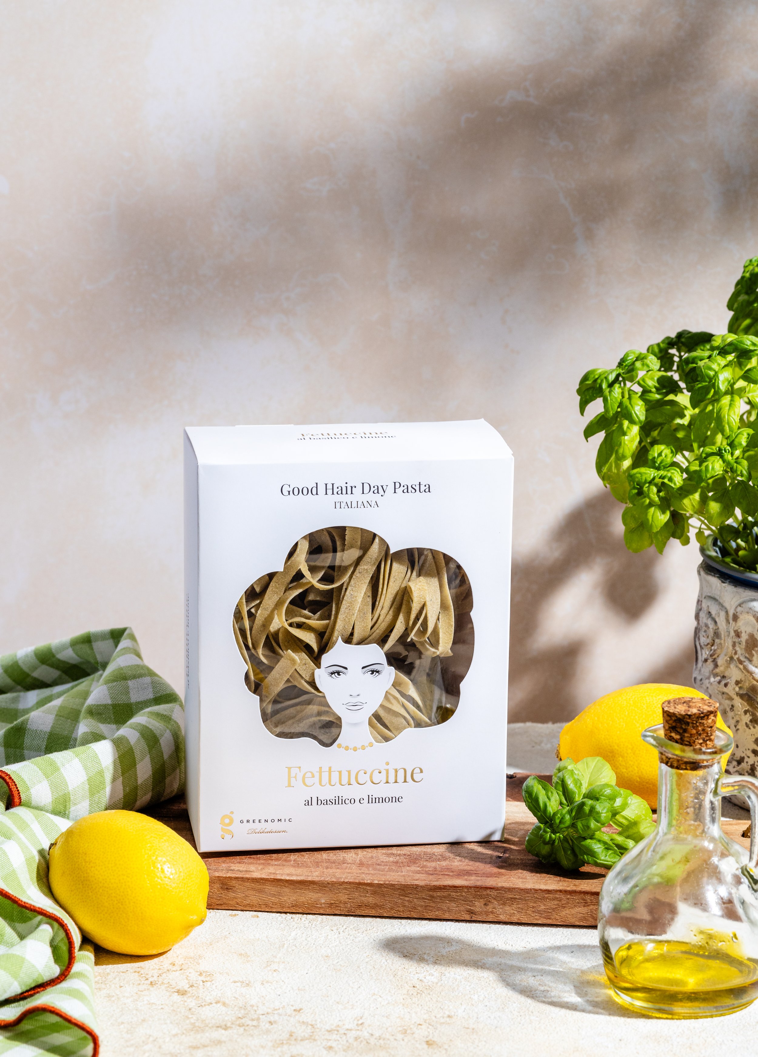 Eine Packung vegetarische Fettuccine Pasta mit Basilikum und Zitrone, umgeben von Zitronen, Basilikumblättern, einem Glas mit Öl, und einem grünen und weißen Stofftuch auf einem Holztisch. Im Hintergrund eine Pflanze.