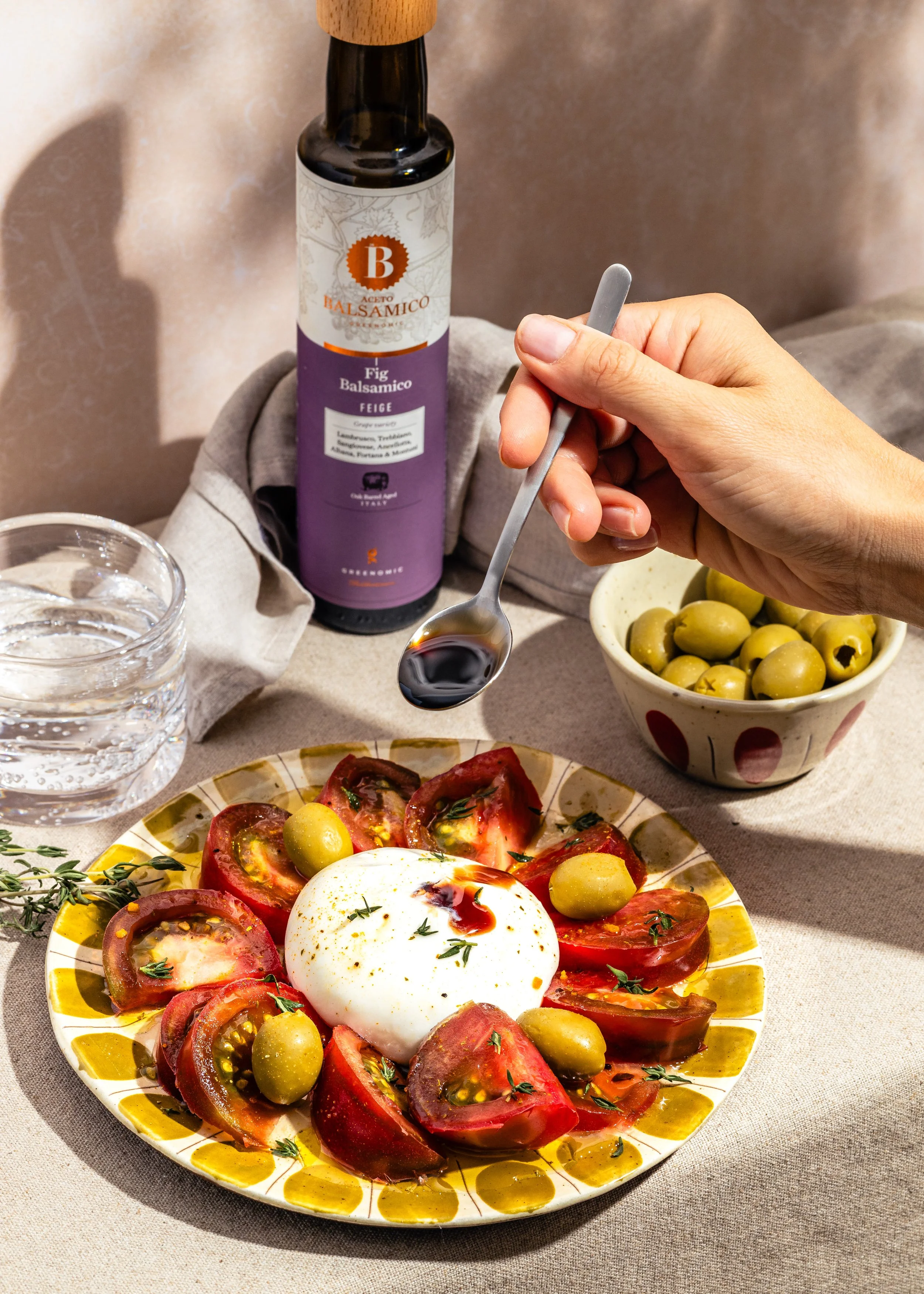 Ein Teller mit Tomaten, Mozzarella, Oliven und einem weichen Ei, darüber Balsamico-Soße, mit Kräutern bestreut, daneben eine Flasche Balsamico-Essig, eine Schüssel mit grünen Oliven und ein Glas Wasser.