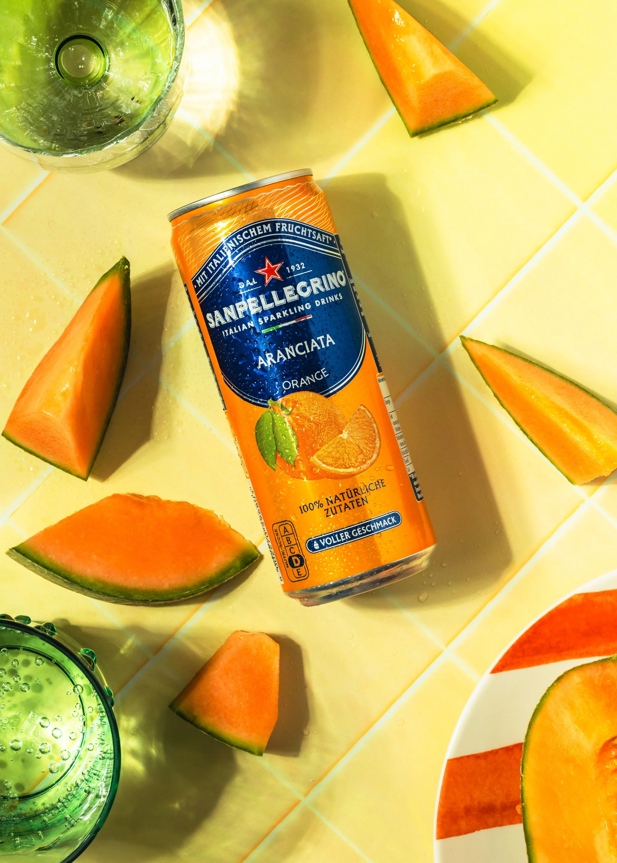 Eine offene Dose Sanpellegrino Aranciata, halb volle grüne Gläser mit Wasser, Melonen- und Papayastücke auf gelbem Fliesenboden.