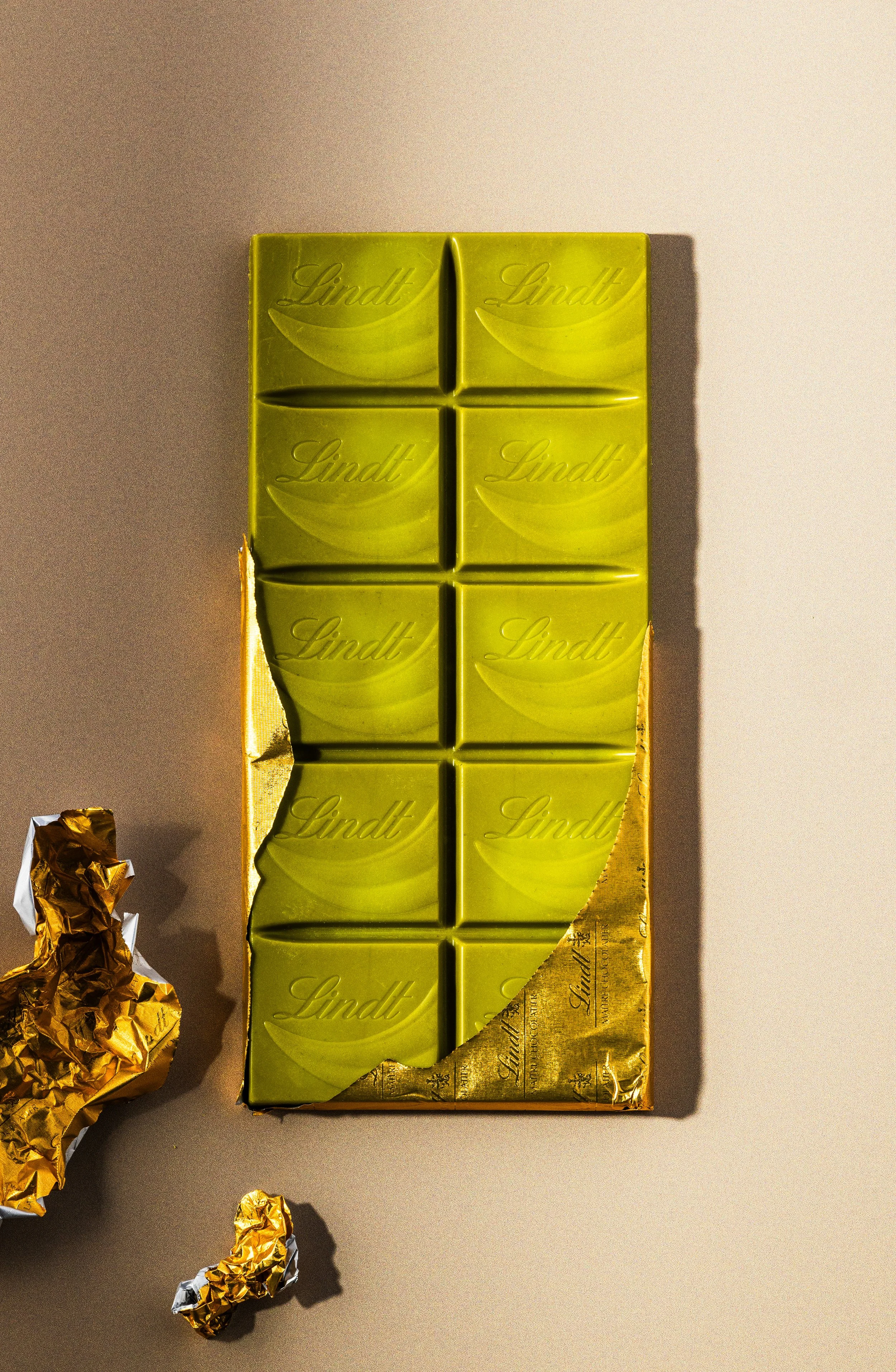 Eine aufgeschnittene grüner Schokolade mit der Aufschrift "Lindt" in Goldfolie, daneben sind zerknitterte Goldfolie und Papierstücke auf einem beige Hintergrund.