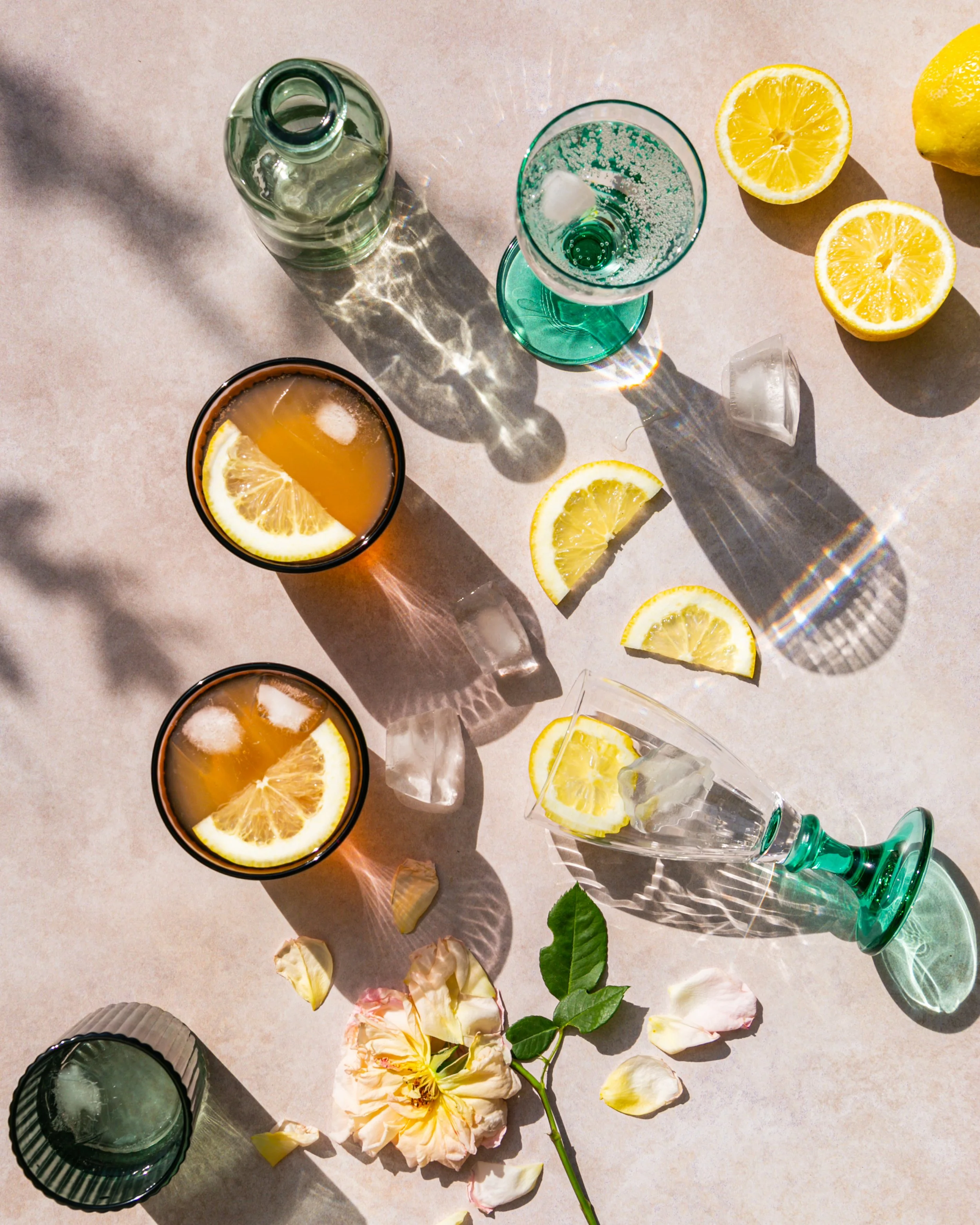 Bilder von zwei halben Zitronen, leeren Gläsern mit Zitronenscheiben, einer Flasche, sowie Blumen und Blätter auf einem hellen Untergrund, alles bei Sonnenlicht.