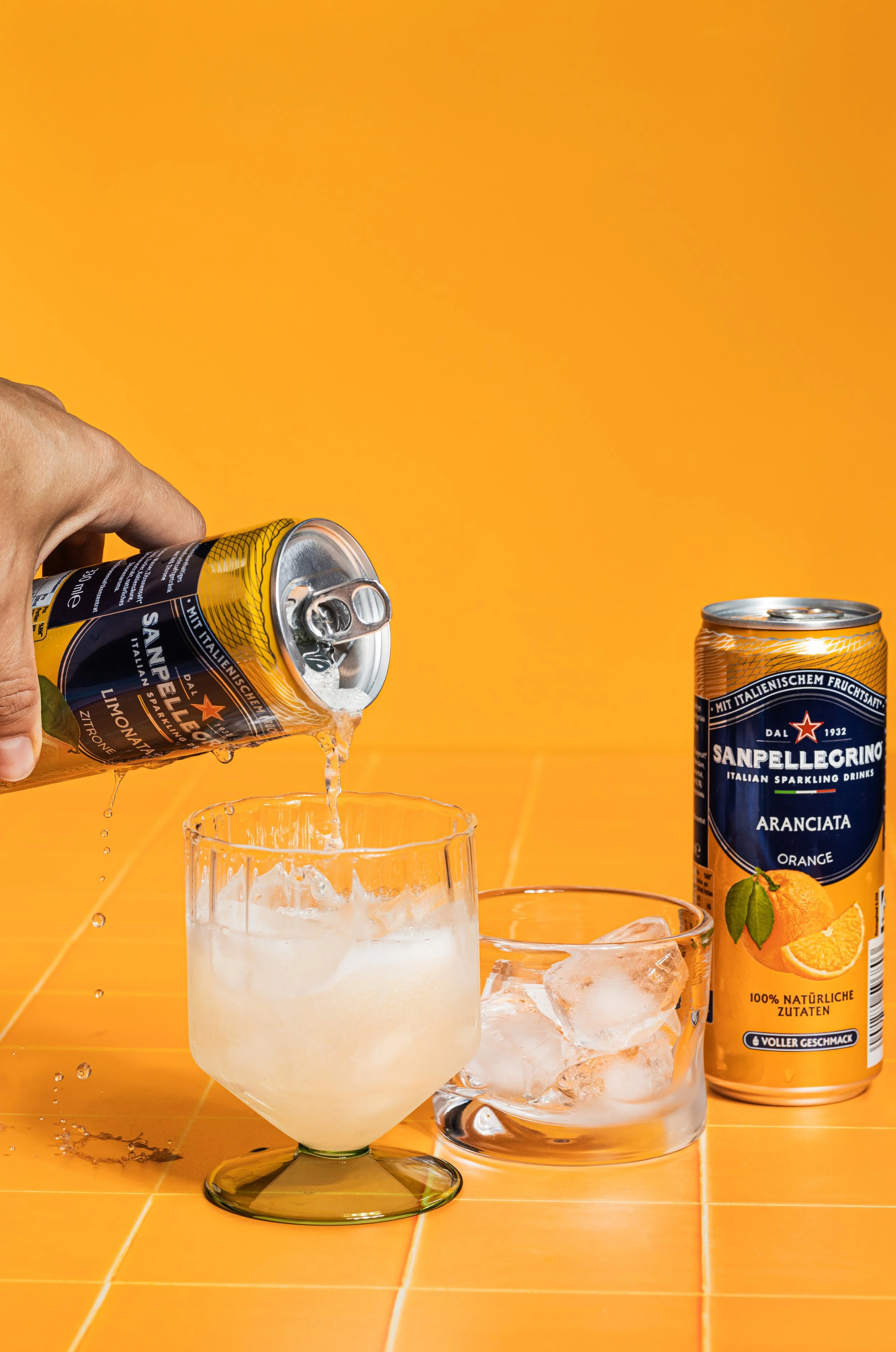 Hand gießt einen Orangensaft in ein Glas mit Eis auf einem orangefarbenen Hintergrund; im Hintergrund eine Dose San Pellegrino Orangenlimonade, ein Glas mit Eis, eine Glasvase mit Wasser und Eis, alles auf einer orangefarbenen Tischplatte.