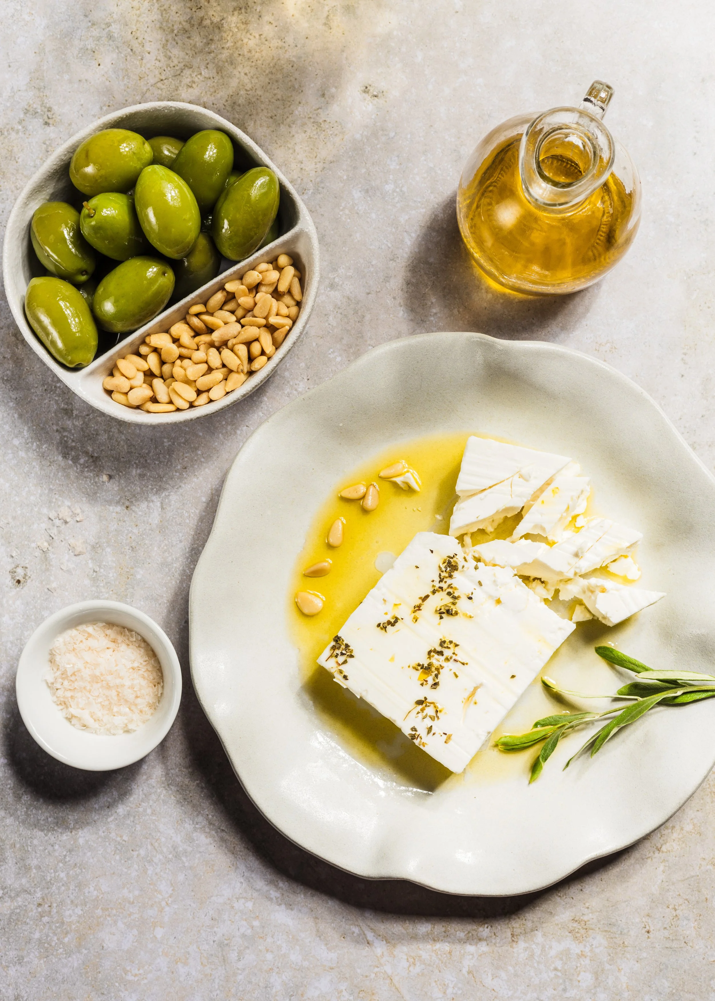 Blick auf eine Platte mit zerbröckeltem Feta-Käse, Olivenöl, frischer Salbeiblatt, sowie ein Schüssel mit Salz, eine Flasche mit Olivenöl, eine Schüssel mit Pinienkernen, grüne Olive und Kerne in einer Schale.