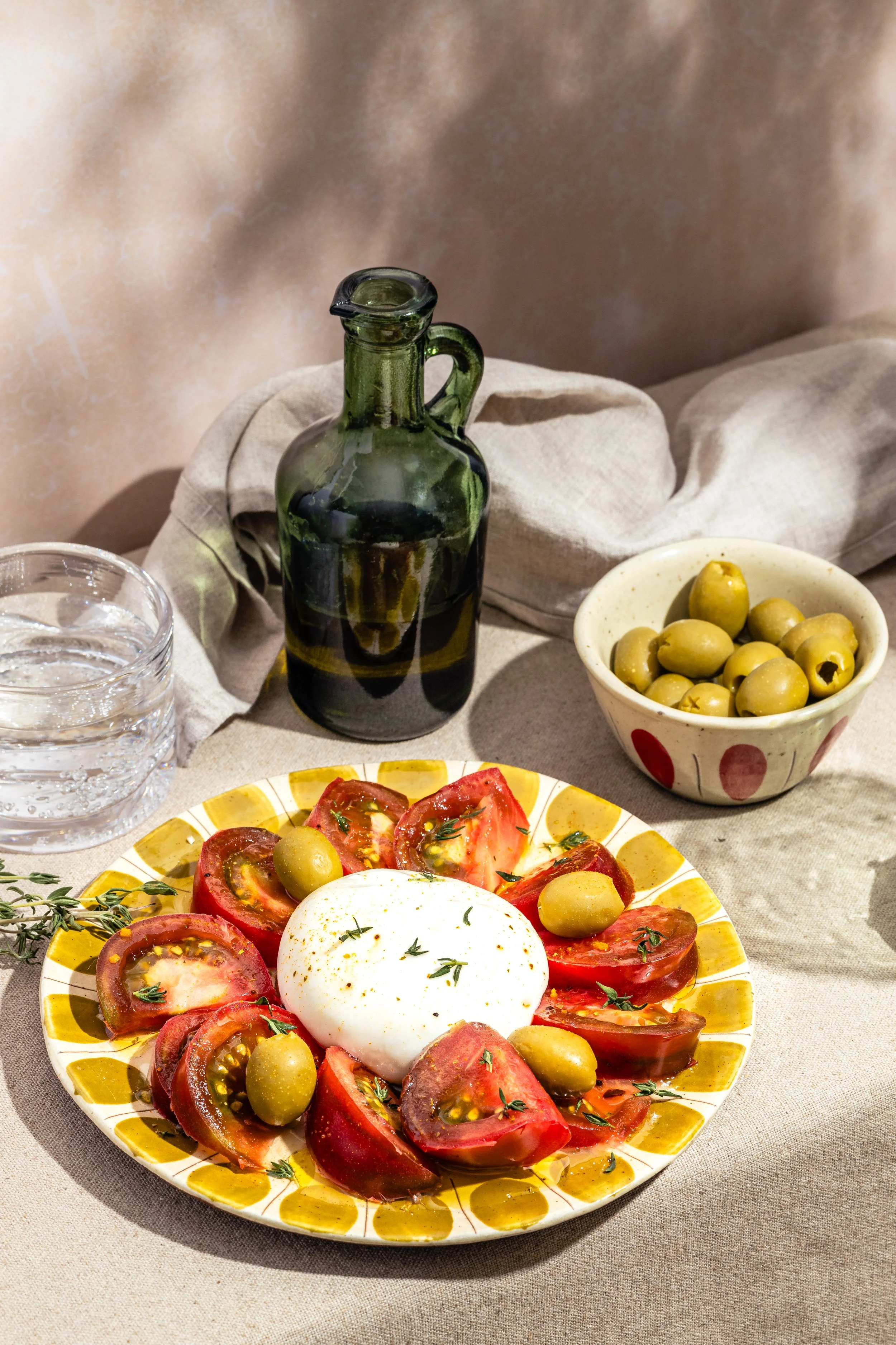 Ein Teller mit Tomaten, grünen Oliven, einem Stück Mozzarella und frischen Kräutern, daneben eine Flasche Olivenöl und eine Schale mit grünen Oliven, auf einem Tisch mit beigeunter Decke.
