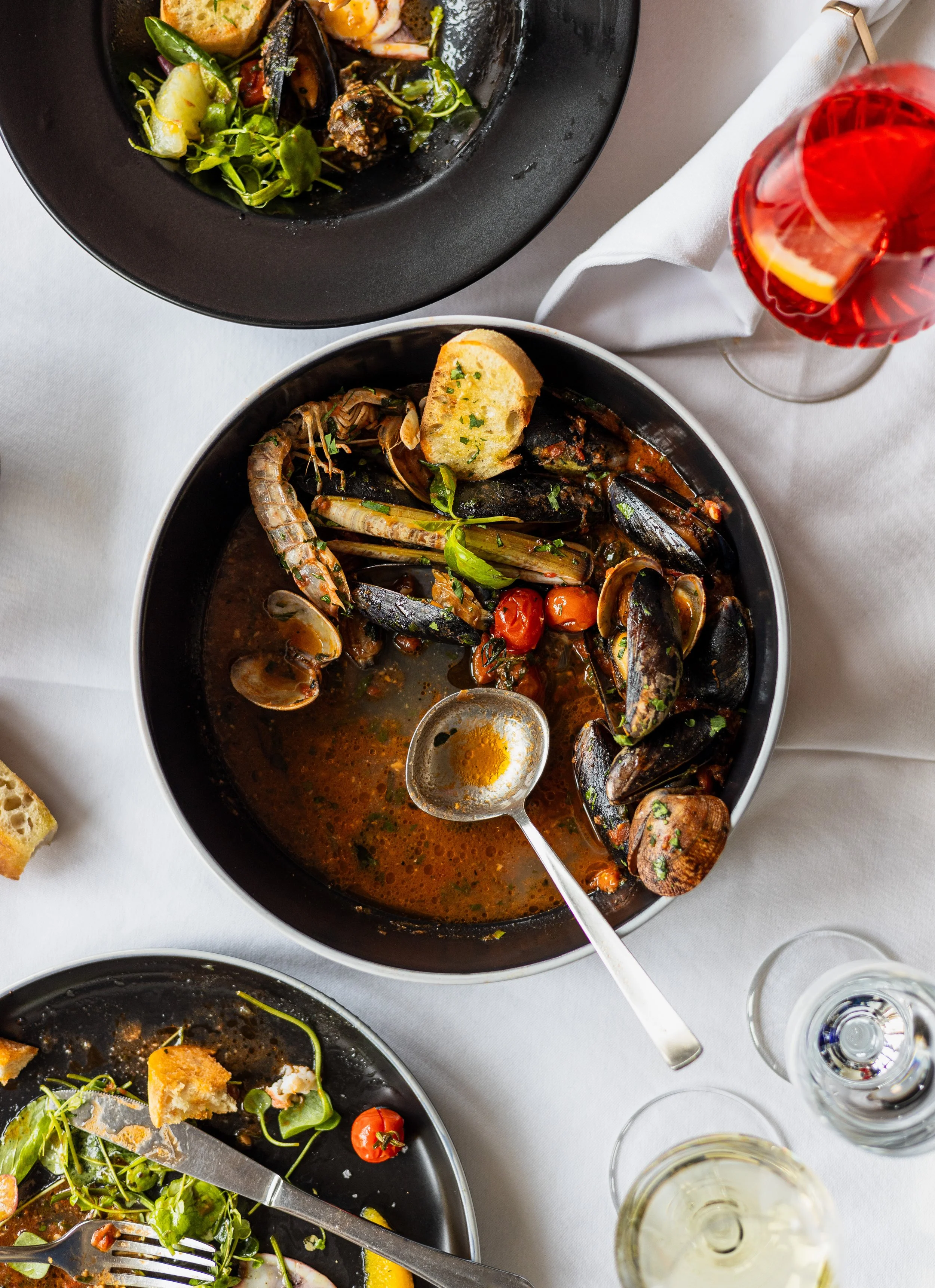Ein Tisch mit mehreren Tellern mit Meeresfrüchten, darunter Muscheln, Garnelen und Fisch in einer braunen Sauce, begleitet von einem Stück knusprigem Brot, sowie Gläser mit weißen und roten Getränken.