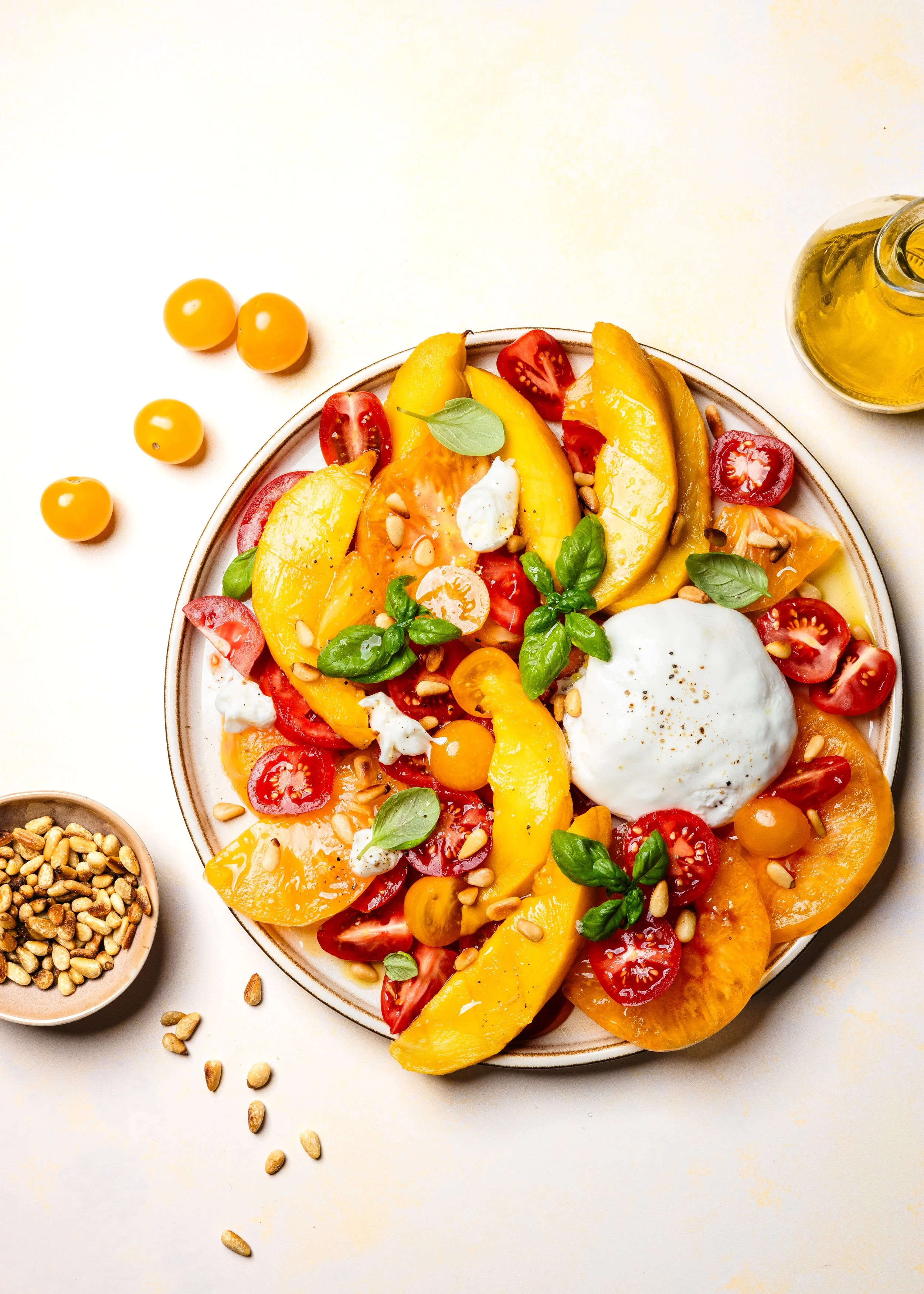 Bunte Caprese-Salat mit Tomaten, Mango, Basilikum, Mozzarella und Sonnenblumenkernen auf einem weißen Teller, daneben eine Flasche Olivenöl und eine Schale mit Sonnenblumenkernen.
