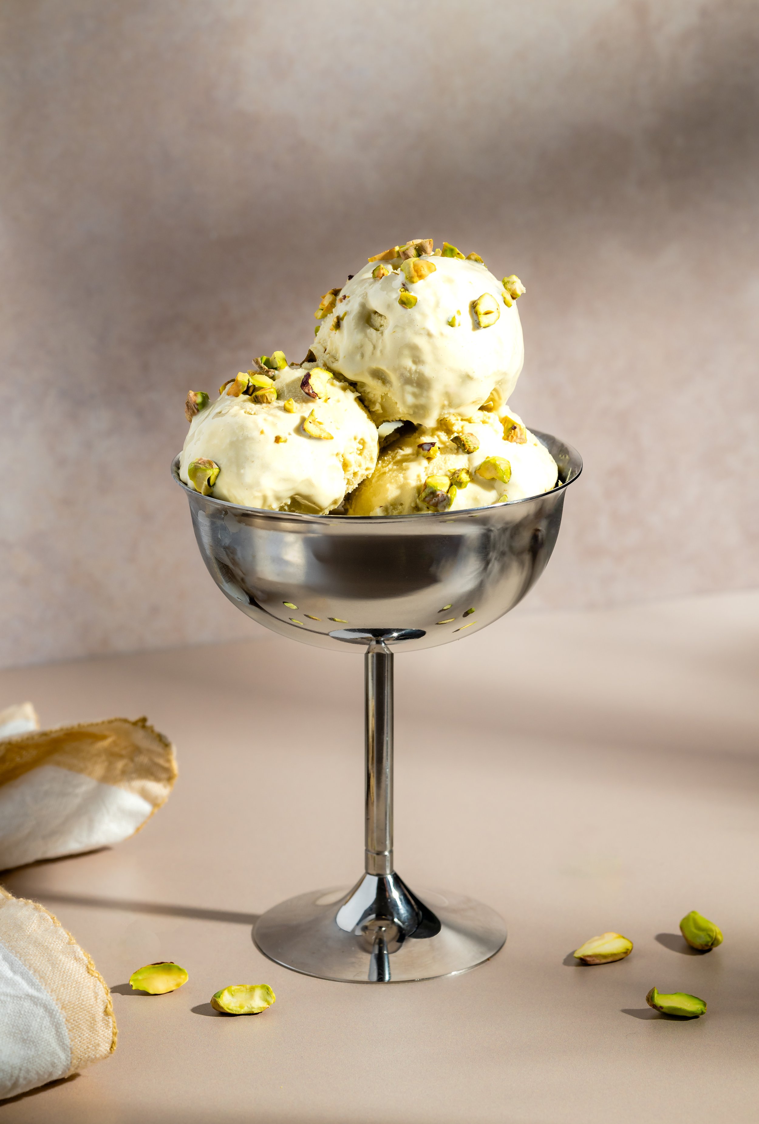 Eisschüsse mit Pistazien in einem silbernen Eisbecher auf einem hellen Hintergrund, einige Pistazien und ein Stück Baiser sind daneben