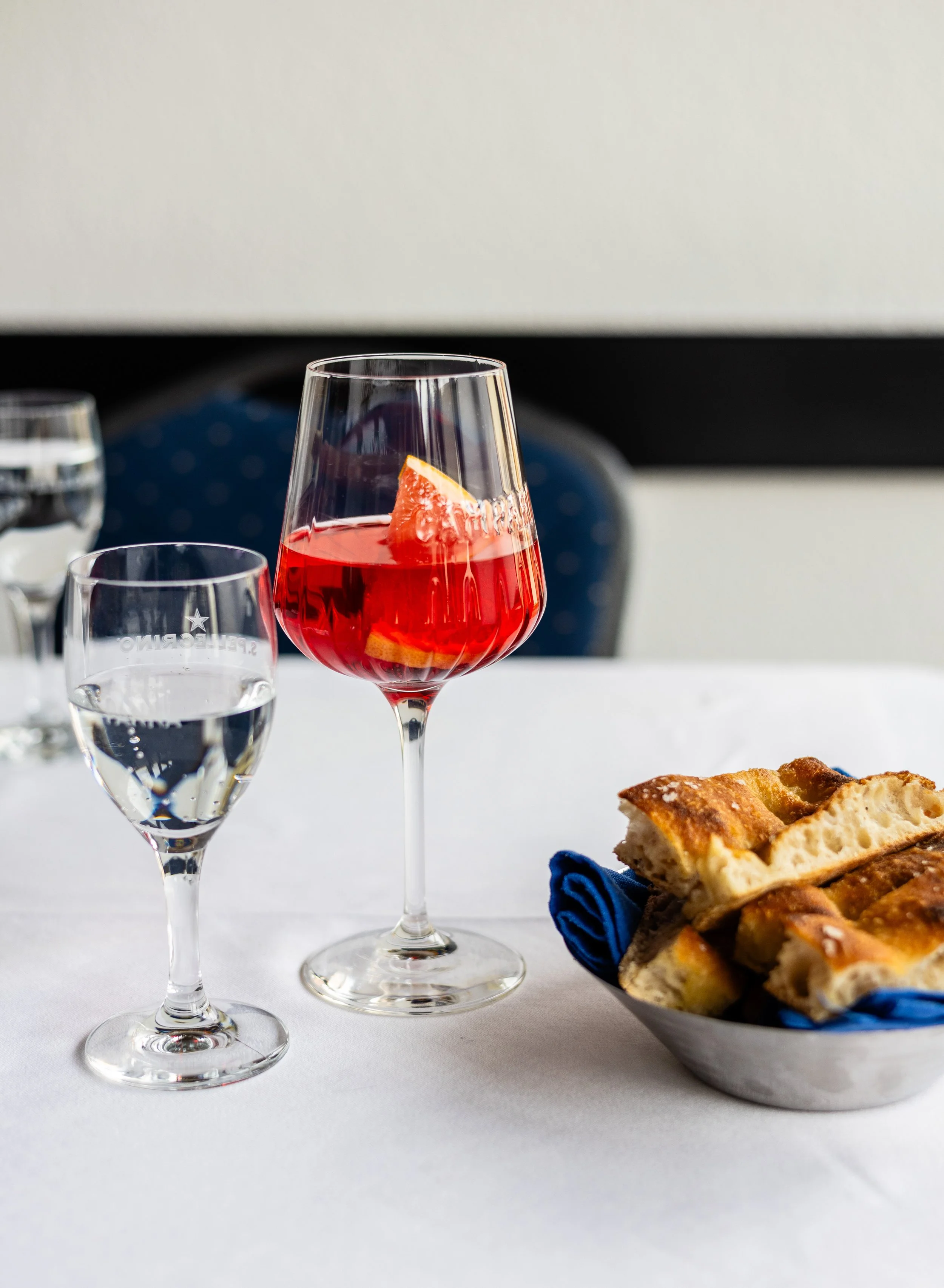 Ein Glas mit rotem Getränk und einer Grapefruit-Scheibe, daneben zwei Wasserhähne und eine Schüssel mit Baguette-Laibchen auf einem weißen Tischtuch.
