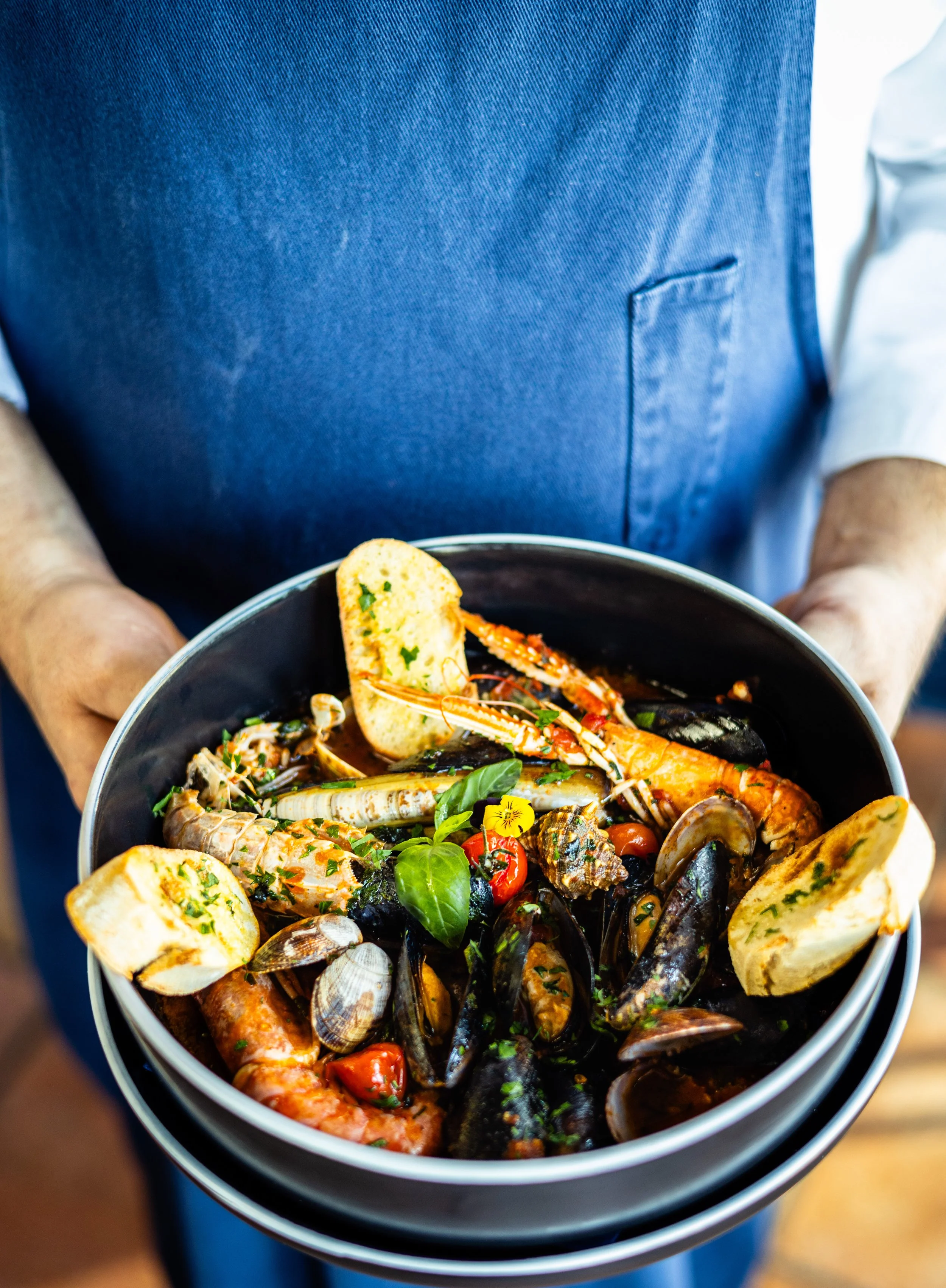 Ein bekannteres Gericht mit Meeresfrüchten in einer Schüssel, mit Garnelen, Muscheln, Kräutern, Brot und Tomaten, serviert von einer Person in blauer Schürze.