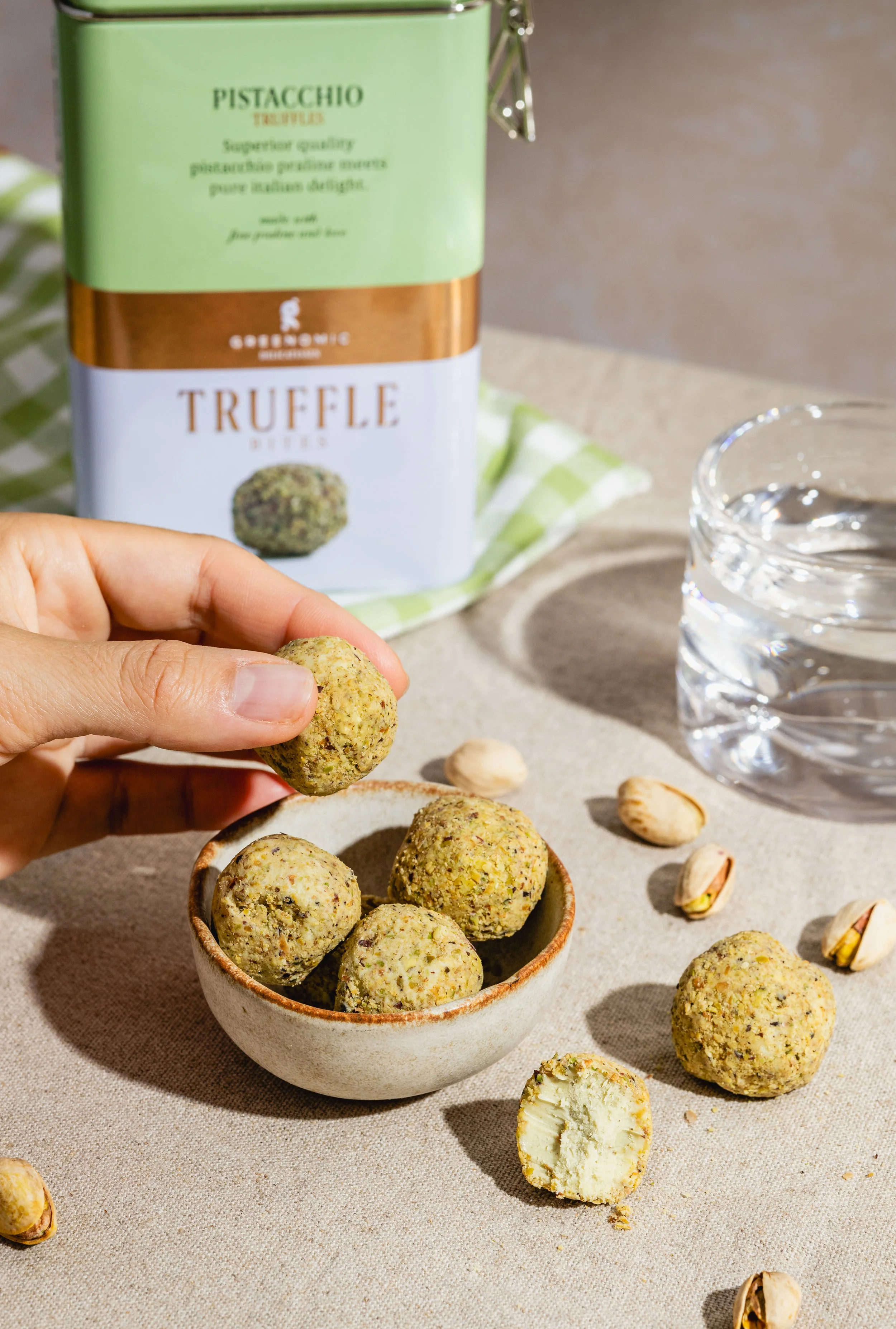 Hand hält eine Pistazien-Protein-Ball, umgeben von weiteren Pistazien-Bällchen in einer Schüssel, im Hintergrund eine Dose Pistazien-Protein.