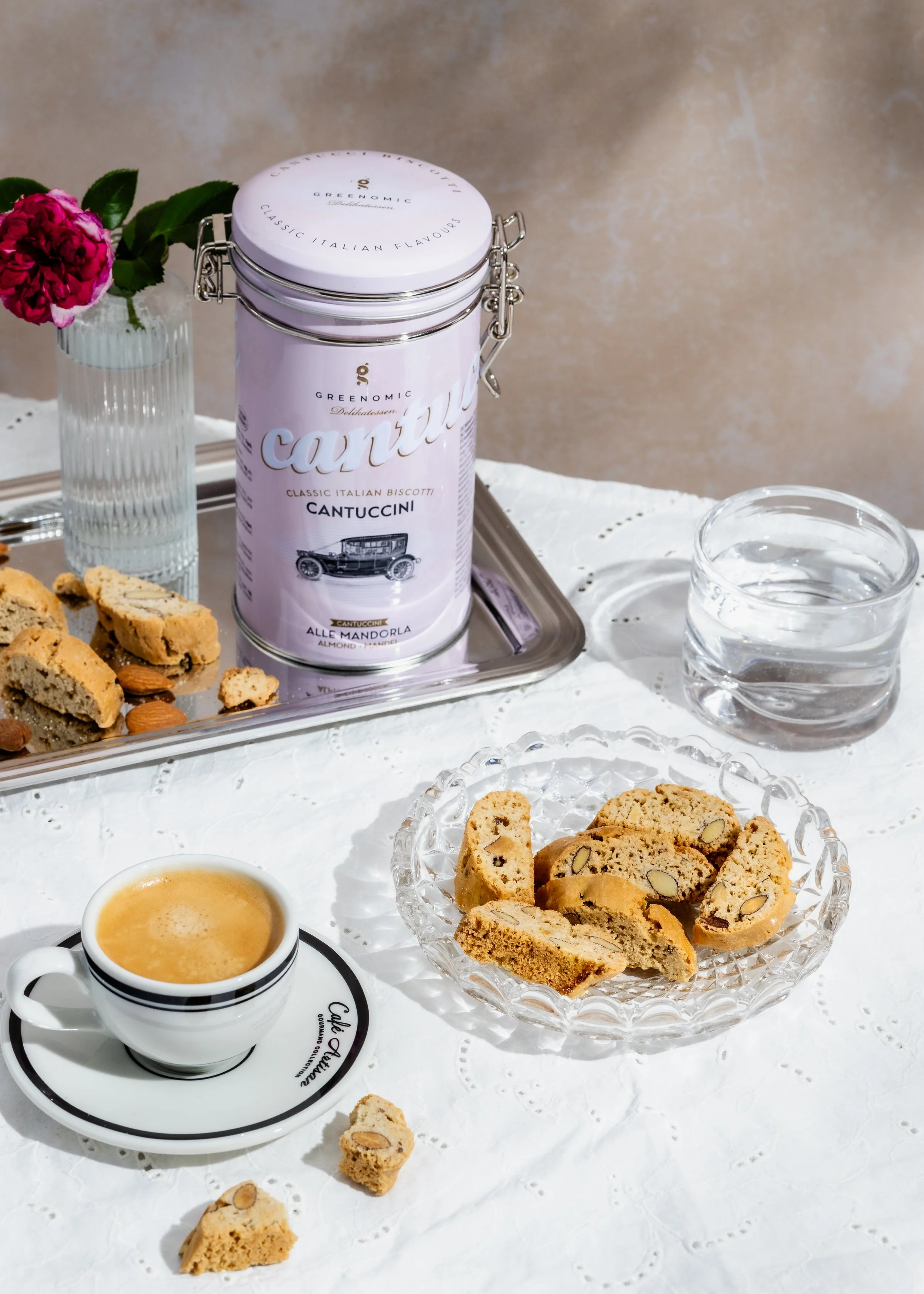 Tasse Kaffee, Kekse, eine Flasche mit Cantuccini-Keksen, eine Vase mit roter Blume, ein Glas Wasser auf weißem Tisch bei Kaffeetafel.