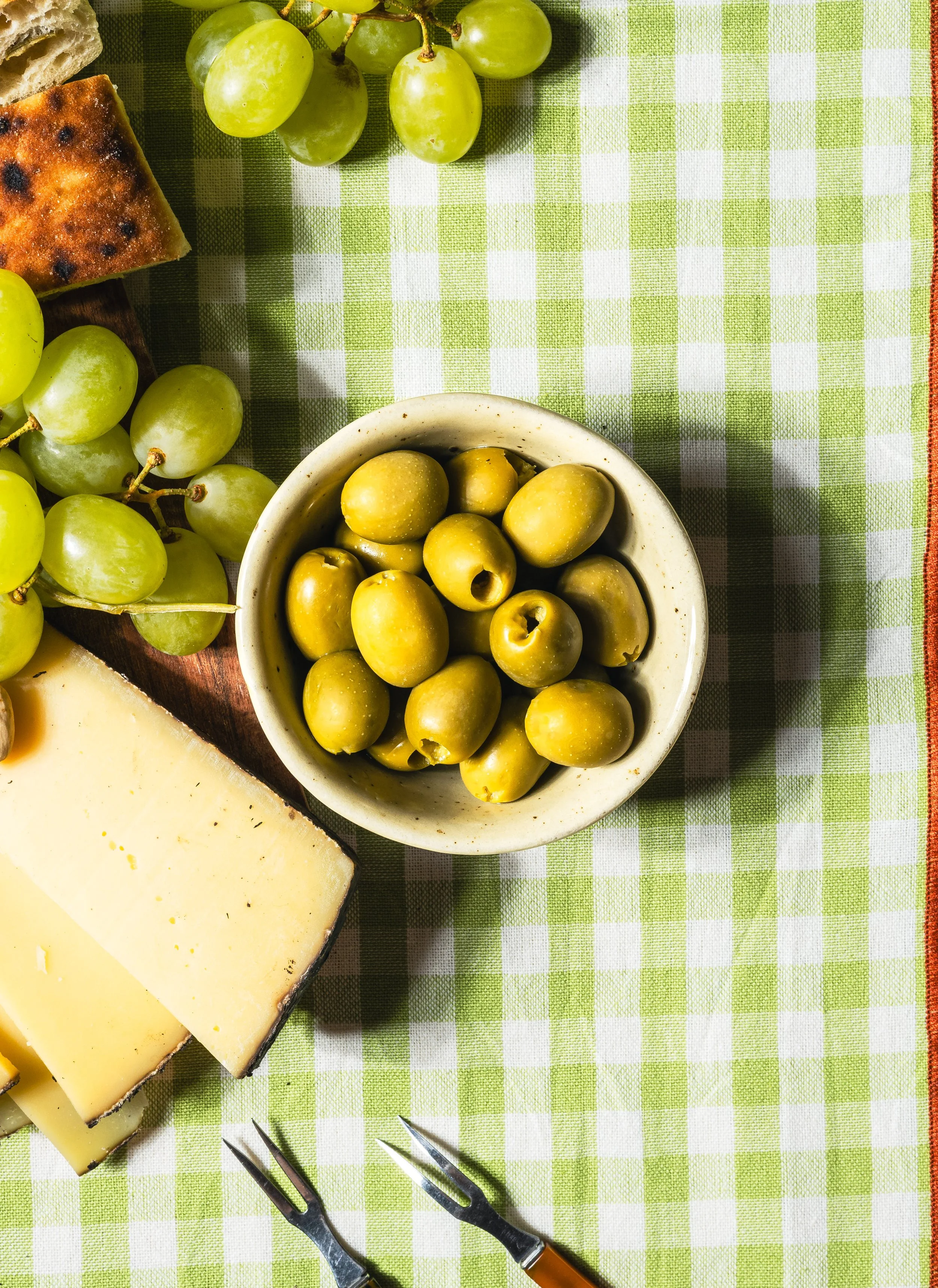 Grüne Trauben, ein Stück Käse, ein Baguette, grüner Oliven, ein Stück Ziegenkäse, zwei Gabeln liegen auf einem grünen und weißen karierten Tuch.