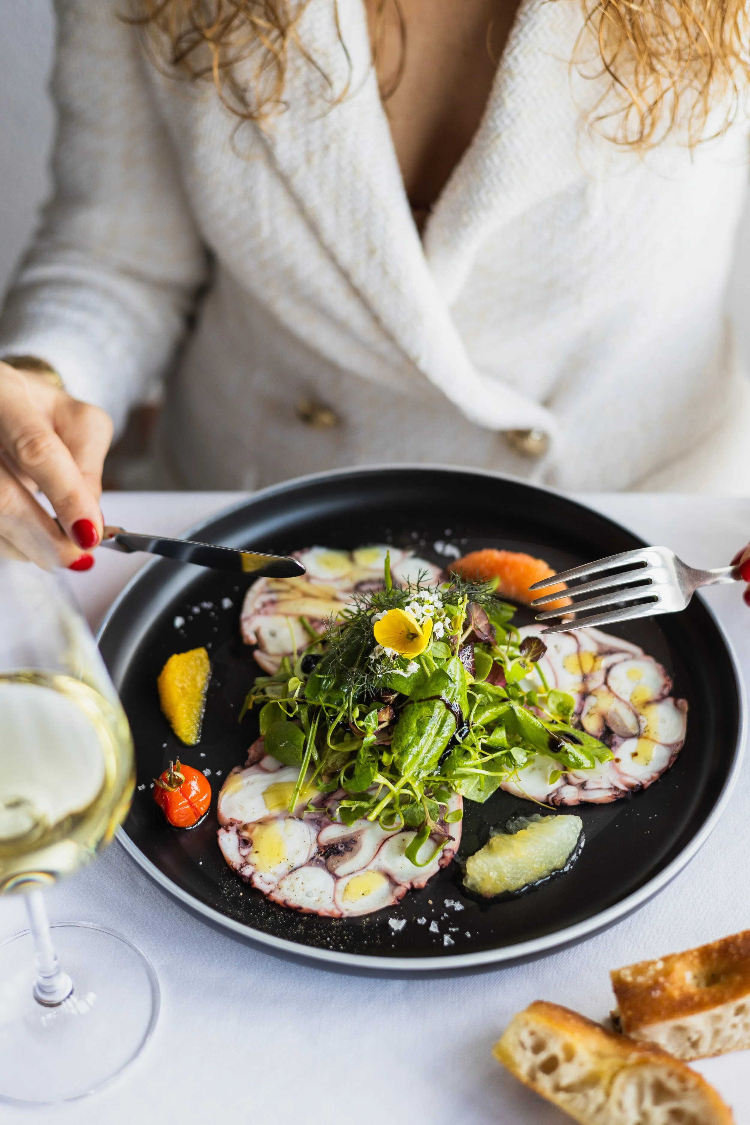 Carpaccio vom mediterranen Oktopus mit Zitrusfrüchten auf einem dunklen Teller, mit einer Hand und Gabel im Bild.