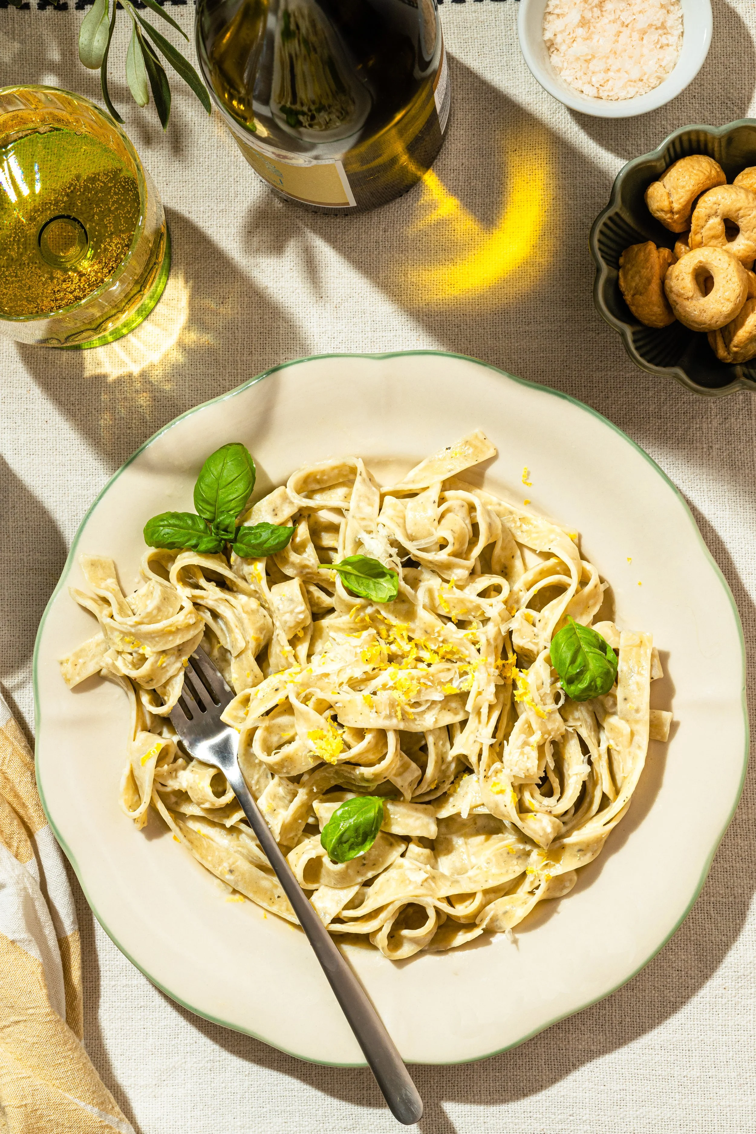 Teller mit Pasta, Basilikum, Zitronenschale, auf einer Tischdecke. Getränke wie Wein oder Bier, Salz, Öl und Kekse sind ebenfalls sichtbar.