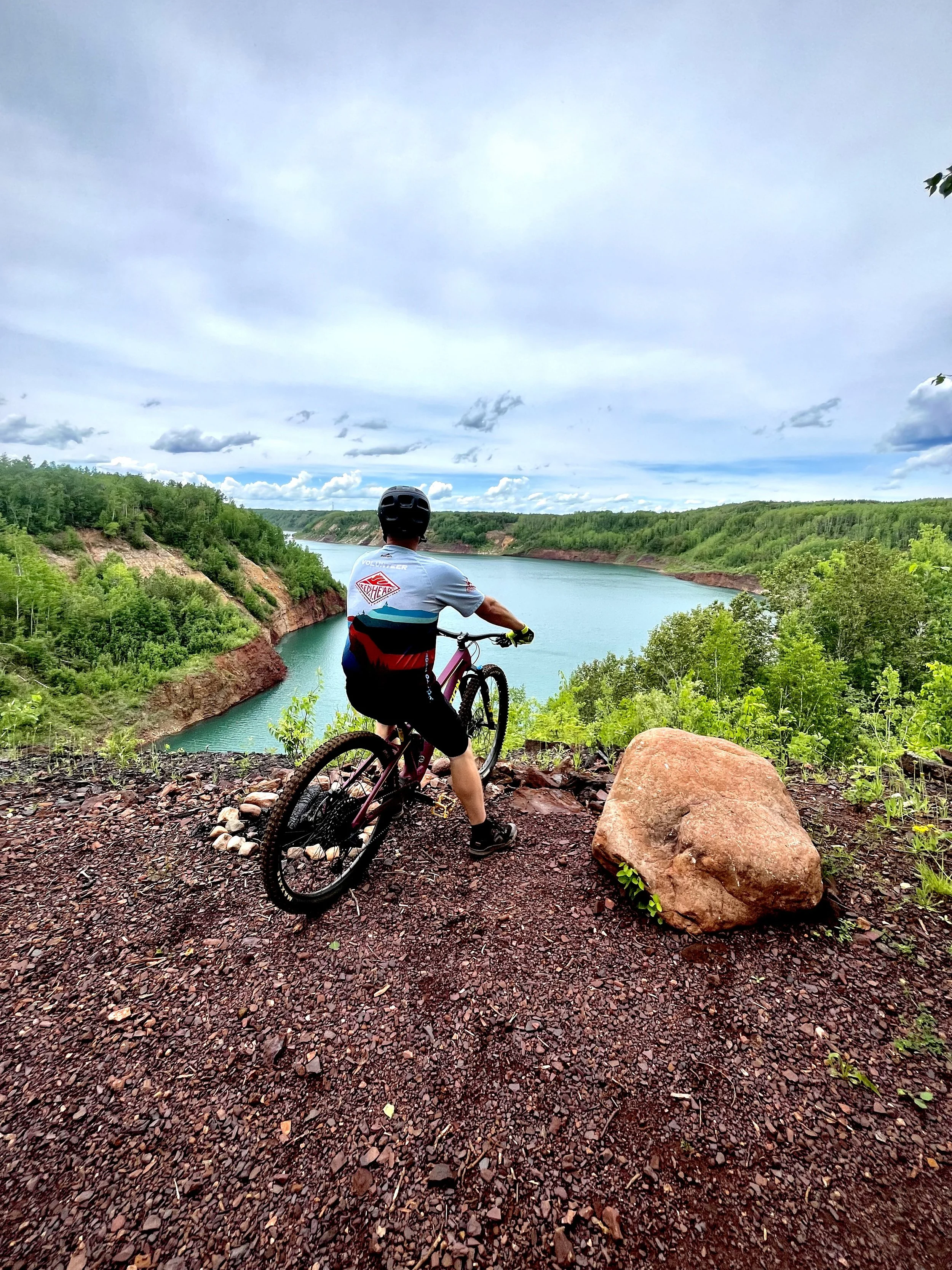 A person with a bicycle sitting on a rocky trail overlooking a river and green hills under a cloudy sky.