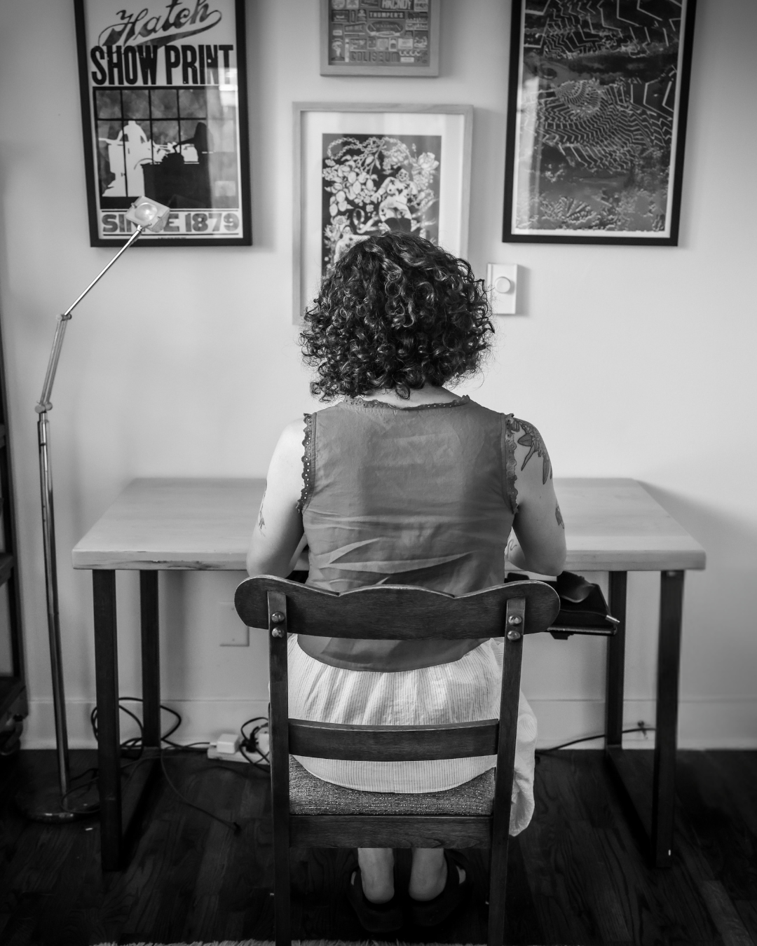 Picture of Vanessa sitting with her back to camera at desk in shirt and skirt with wooden desk and paintings on wall