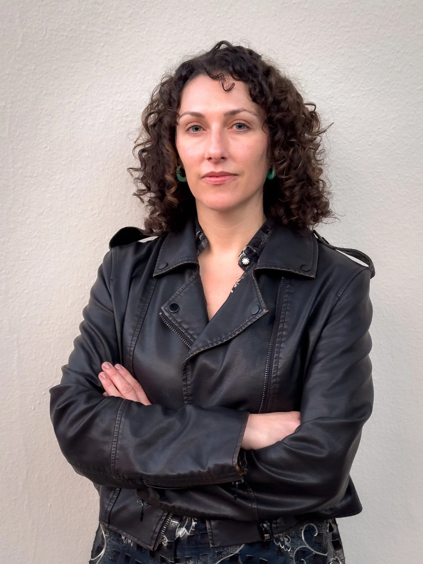 Vanessa with brown curly hair and green earrings with a black leather jacket and crossing her arms across chest