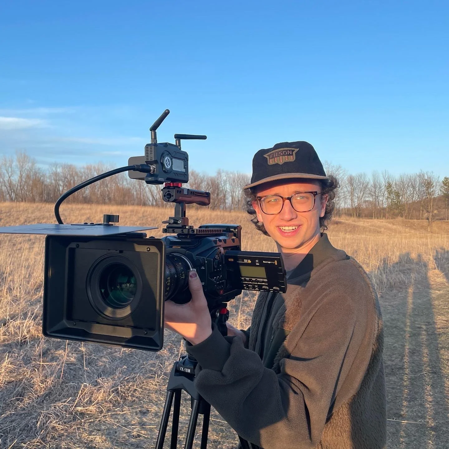 Young man with glasses and a black cap operating a professional video camera outdoors in a field during sunset.