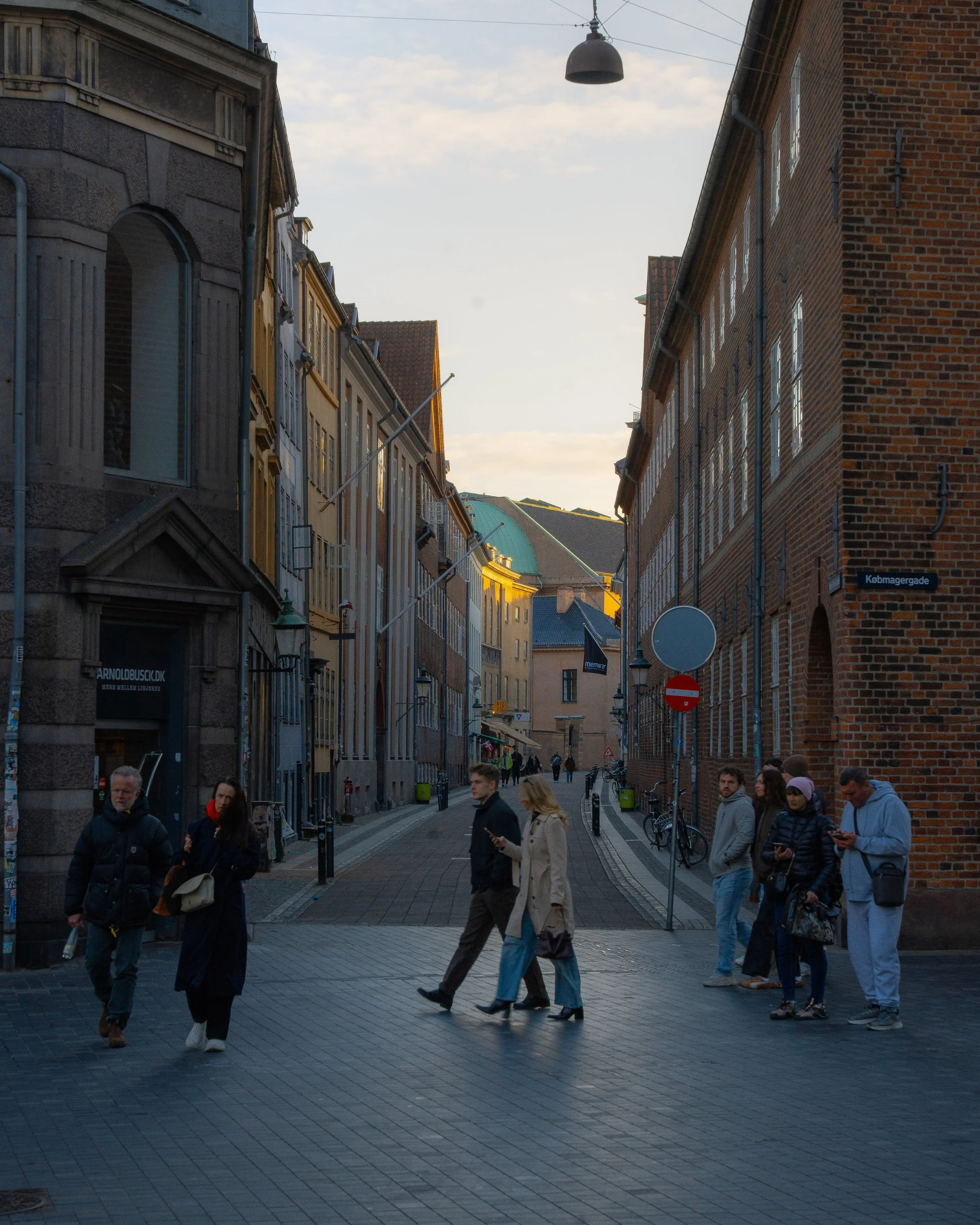 Street scene with people walking on a pedestrian street lined with buildings, some with brick facades, under a partly cloudy sky.