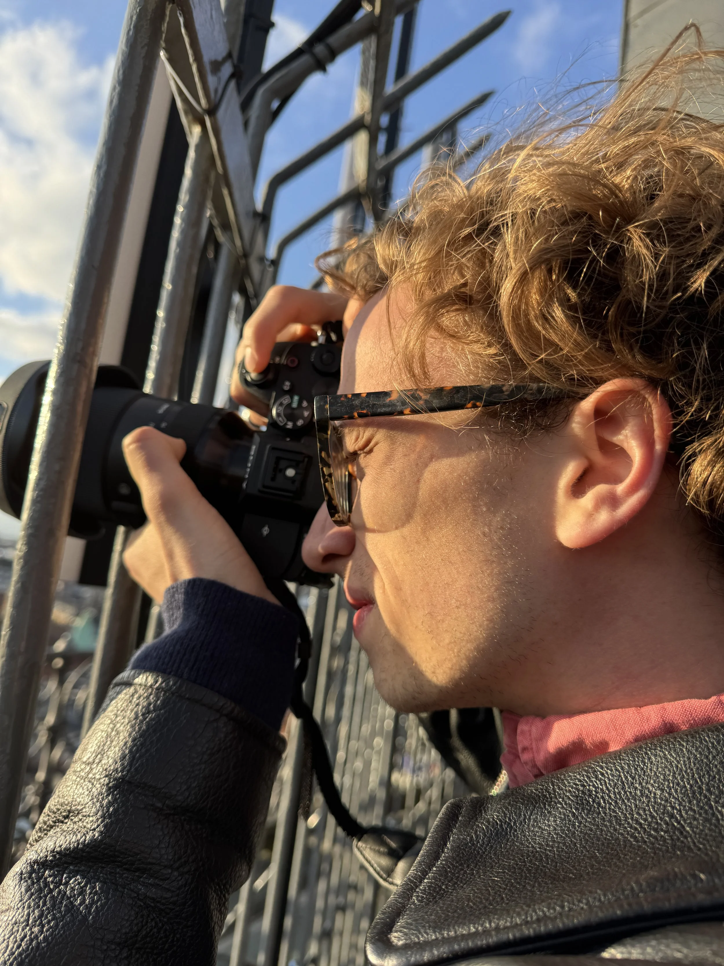 A person with curly hair is taking a picture with a camera, wearing glasses and a black leather jacket, outdoors.