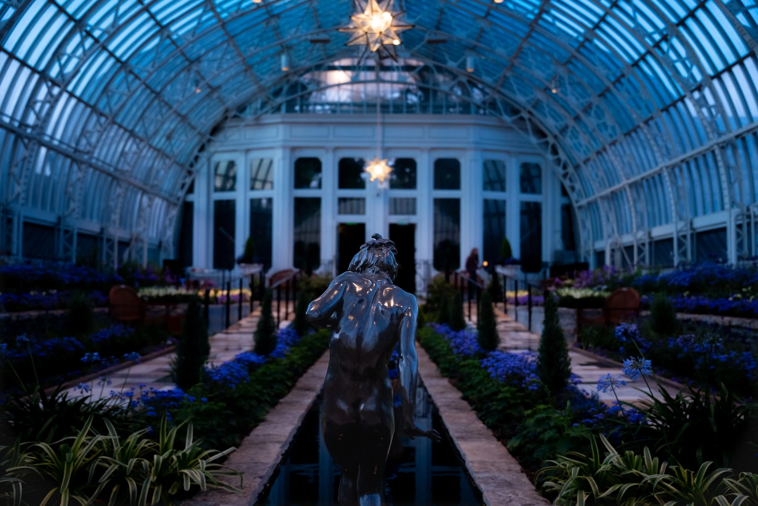 Inside a glass conservatory, a statue of a woman is in the foreground, with a pathway lined with purple flowers leading to large windows and a domed ceiling decorated with hanging star-shaped lights.