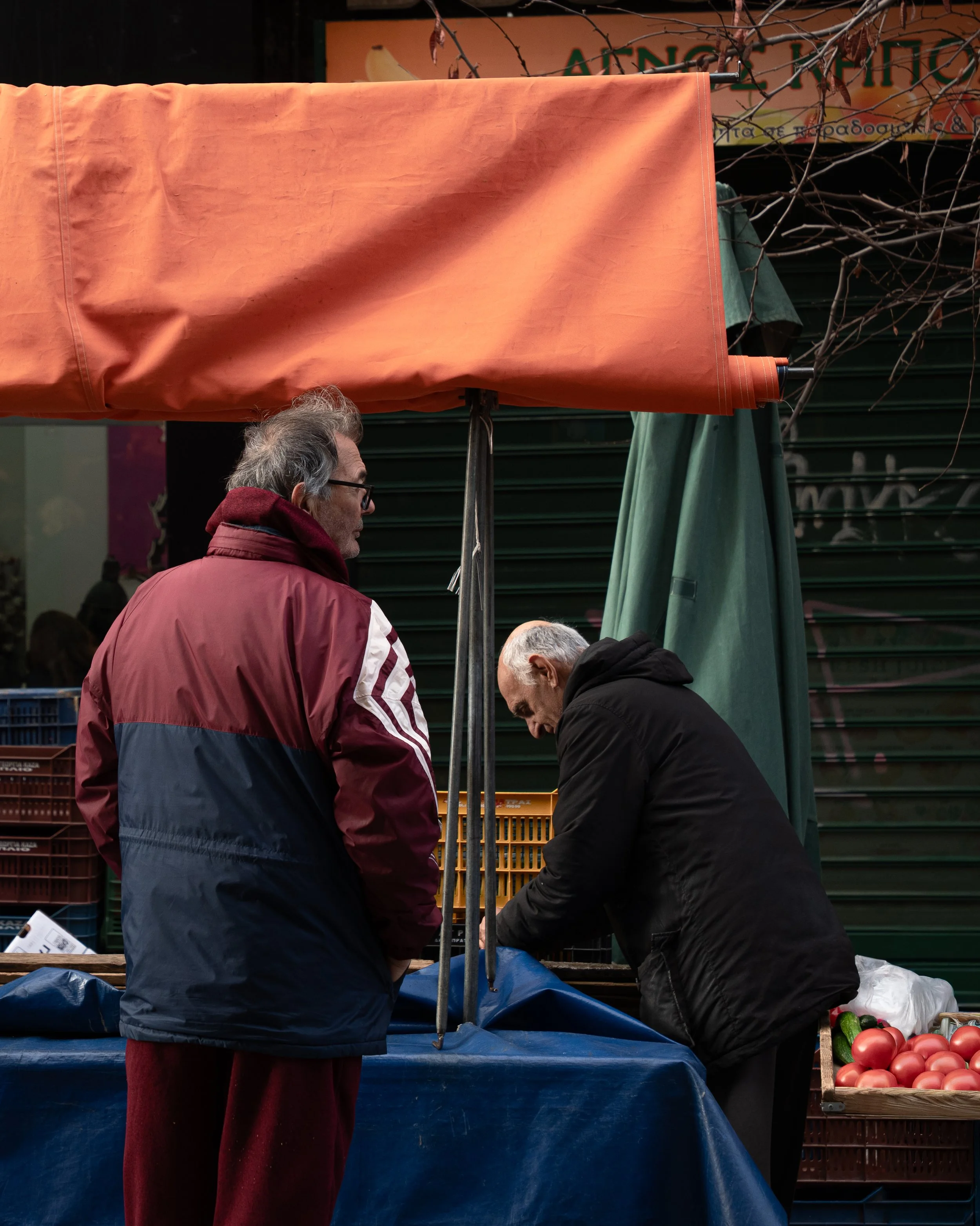 A man in a red and blue jacket and glasses stands at an outdoor market stall with an orange tarp. Two other men are bent over a table under the tarp, organizing produce. There are crates of tomatoes and cucumbers visible to the right. A green umbrell