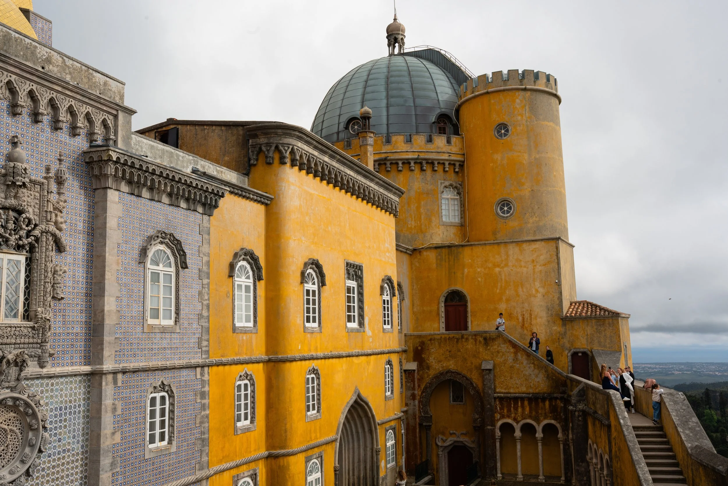 View of a yellow and intricate castle with a large blue-green dome and a tower, with tourists sitting on the steps outside.