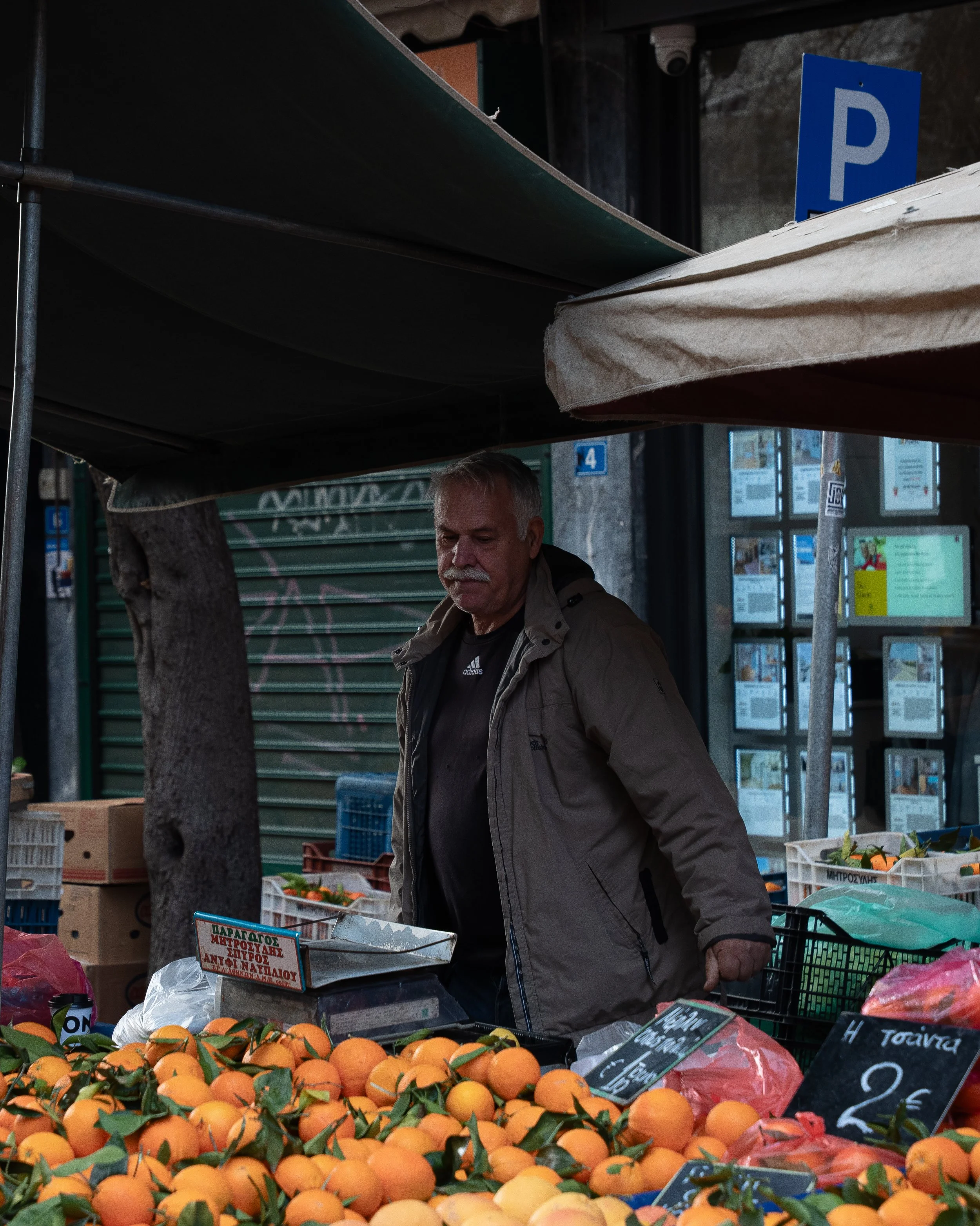 A man shopping at an outdoor market with fresh oranges on display, signs with pricing, and market stalls in the background.